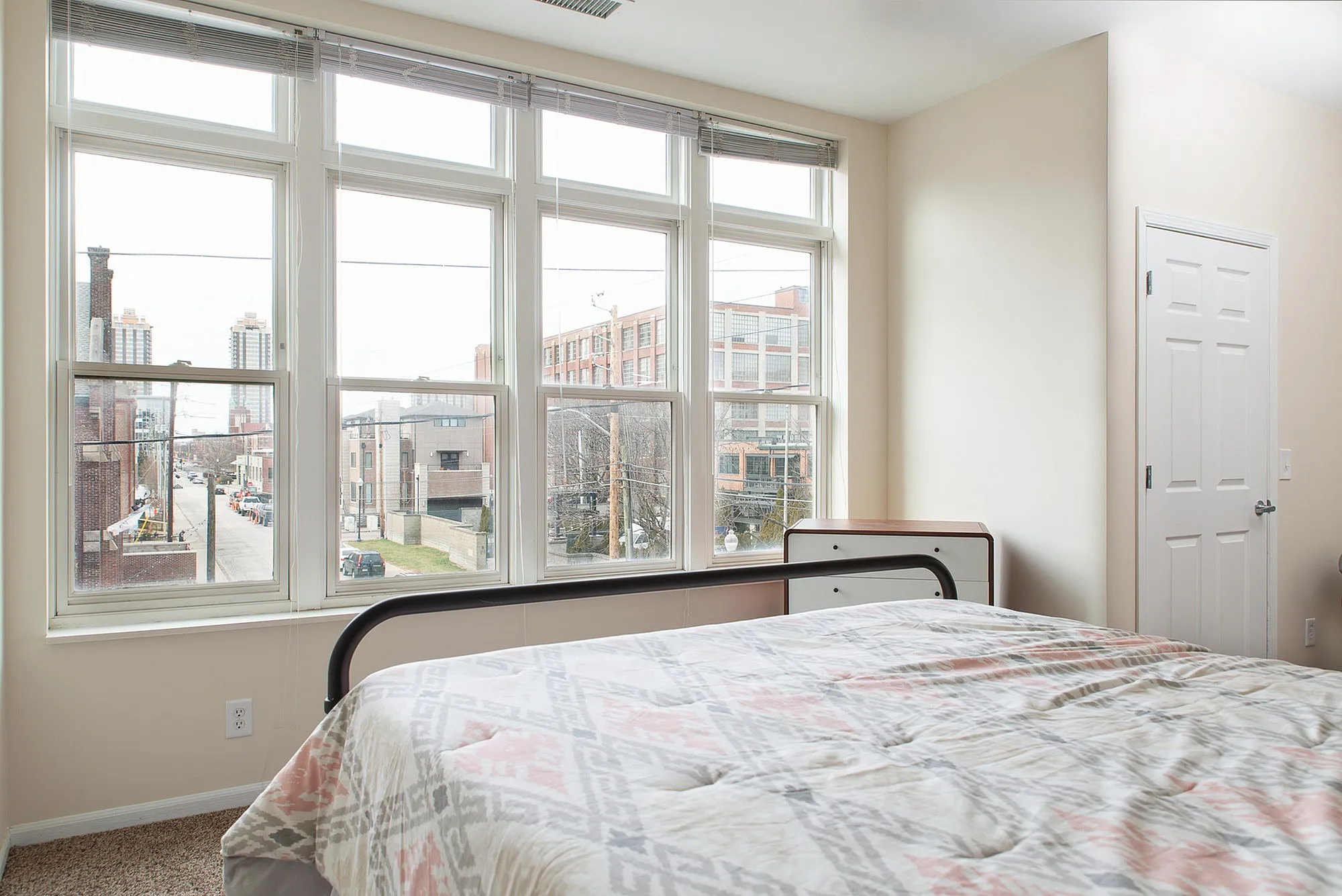 Bright bedroom with large windows offering an urban view, showcasing a cozy, inviting atmosphere.