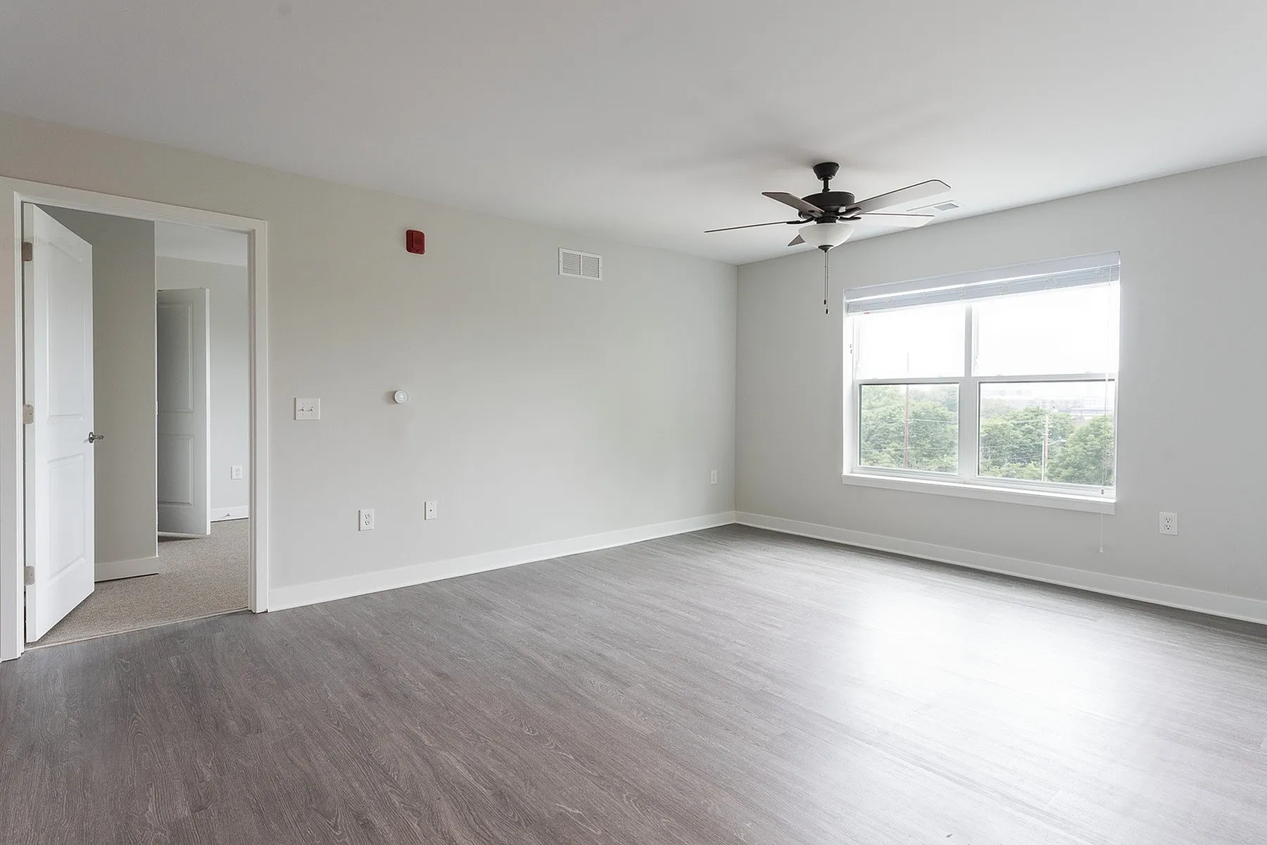 Bright and spacious living room with sleek hardwood flooring, large window, and modern ceiling fan, offering a fresh and inviting ambiance.