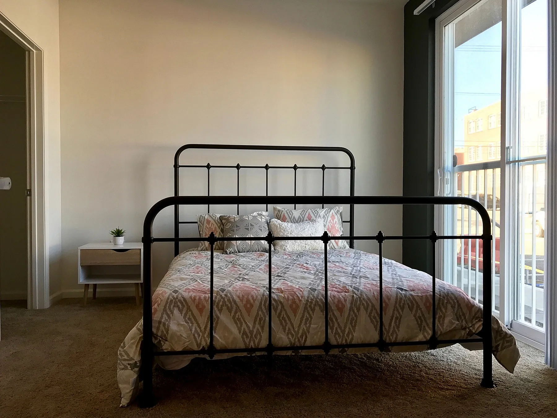 Bright and welcoming bedroom with stylish iron bed frame, patterned bedding, plush carpet, and natural light streaming through large sliding glass doors. Ideal for modern living.