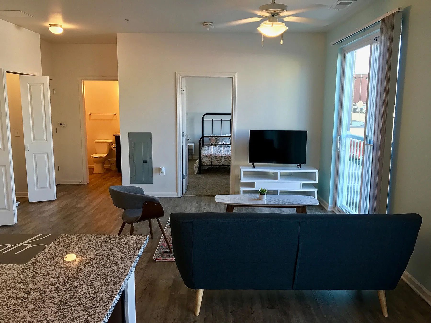 Bright modern living space featuring an open-concept design, sleek wood flooring, and a large window balcony access, leading to a cozy bedroom with ensuite bath.