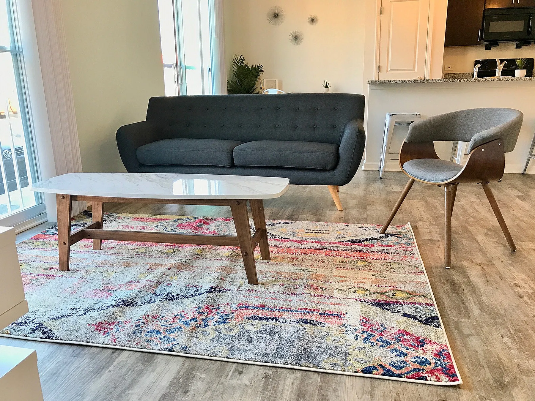 Modern living room with a sleek gray sofa, stylish armchair, and vibrant multicolored rug, complemented by natural light and contemporary decor.