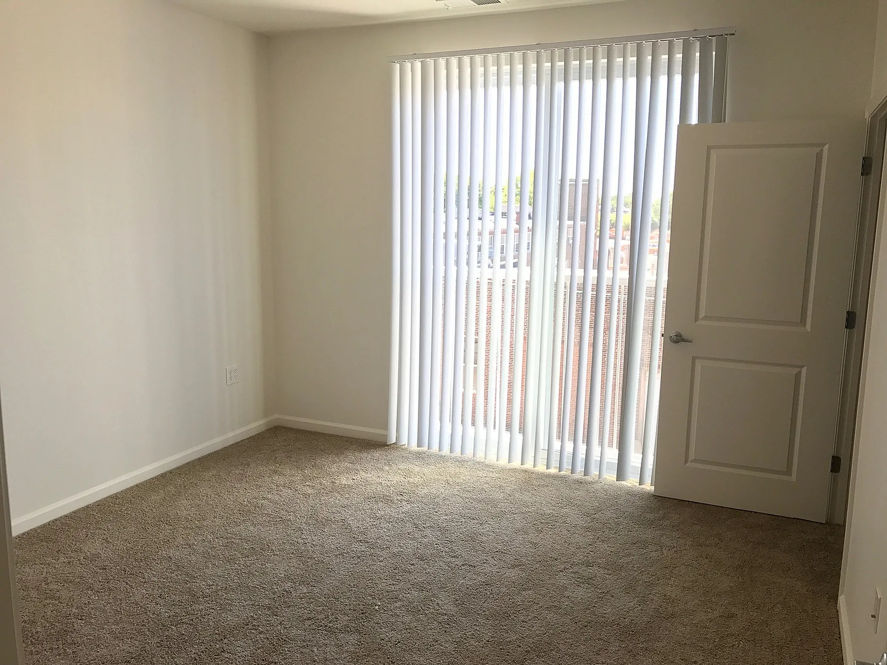 Bright and inviting bedroom with plush carpeting and a large window featuring vertical blinds, offering a peaceful city view.