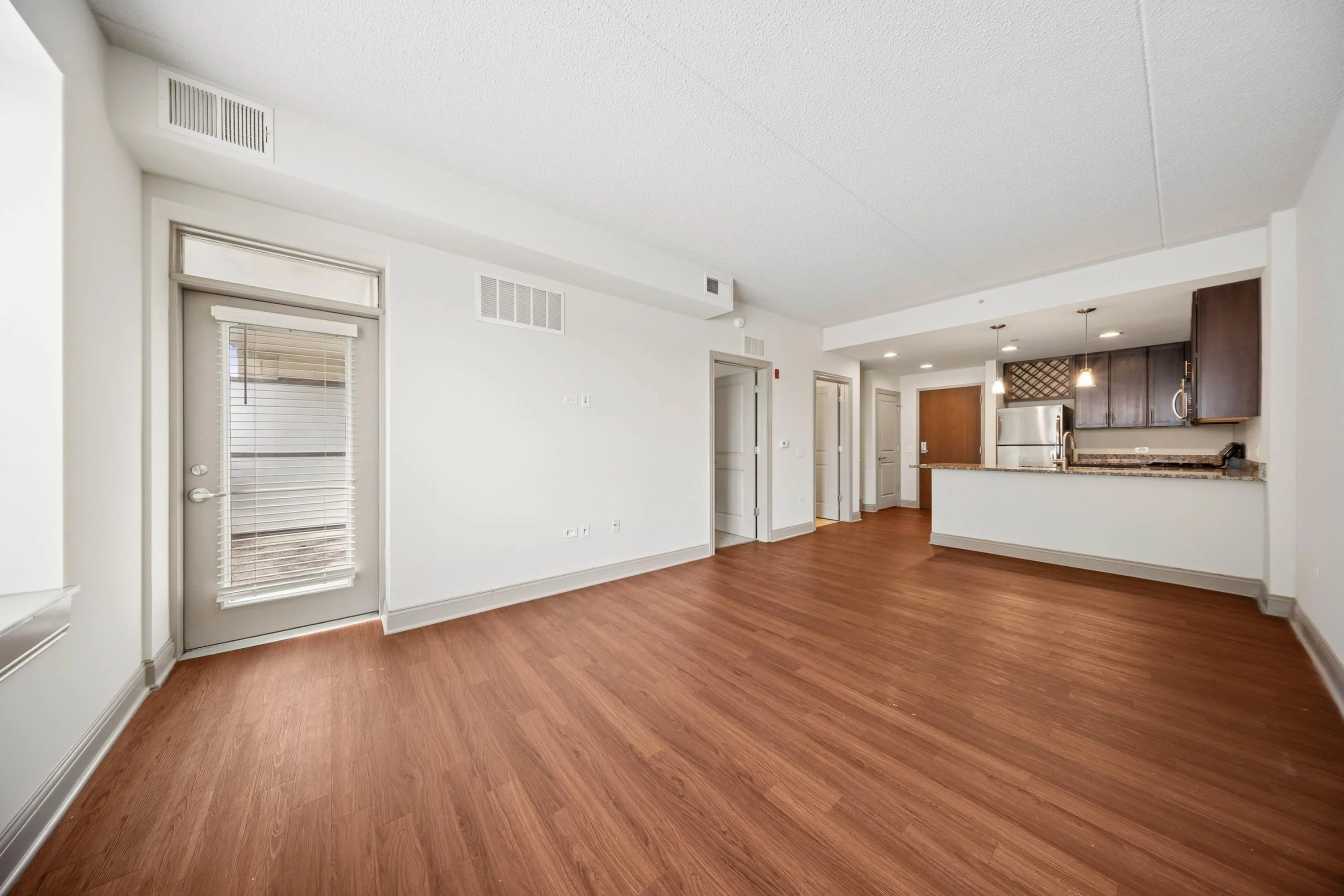 Contemporary open-plan living area featuring sleek hardwood floors, modern kitchen with stainless steel appliances, and abundant natural light from a large doorway.