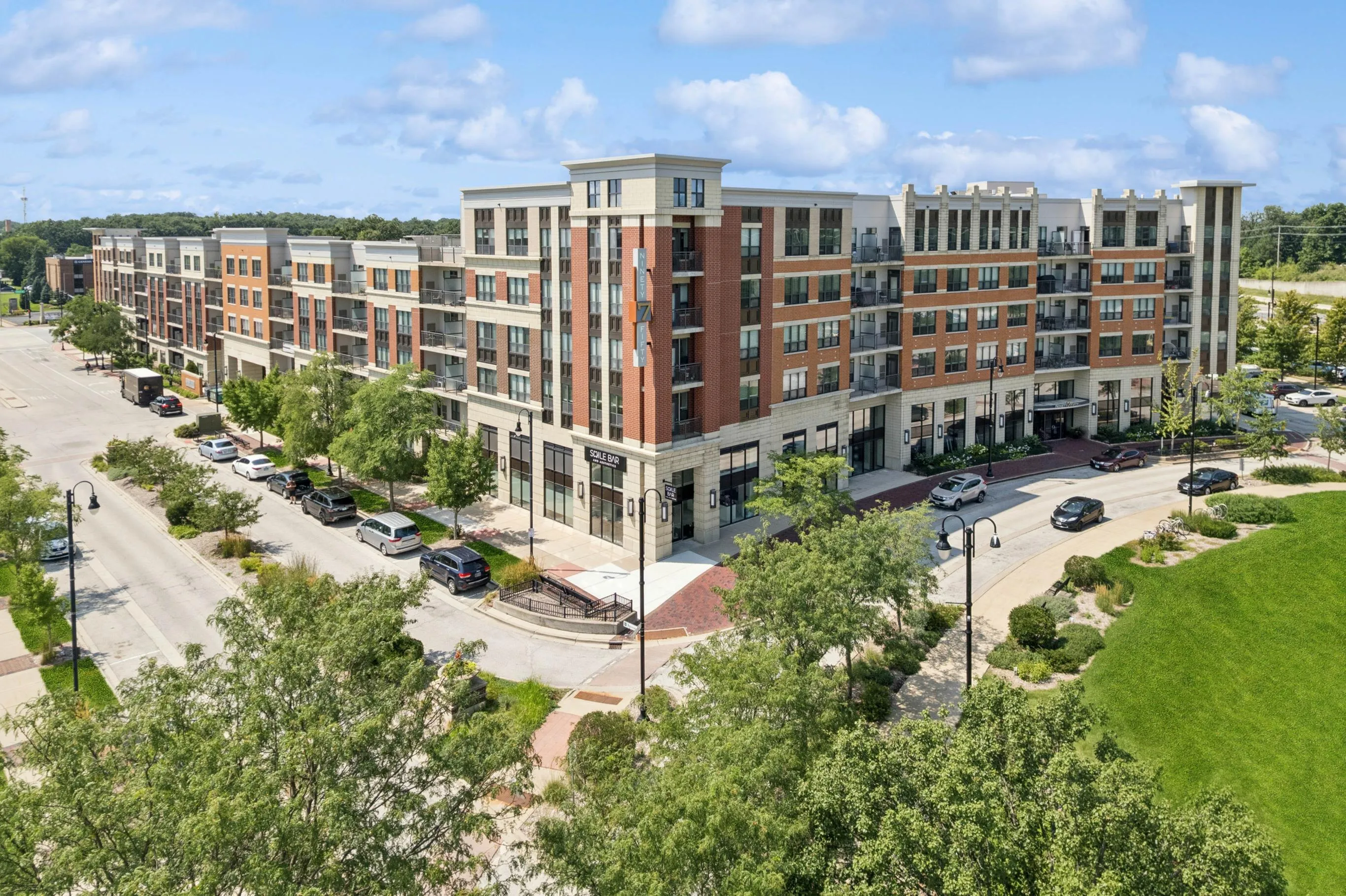 Modern apartment complex featuring stylish architecture with a blend of brick and stone exteriors, set in a vibrant urban environment surrounded by lush greenery and ample parking.