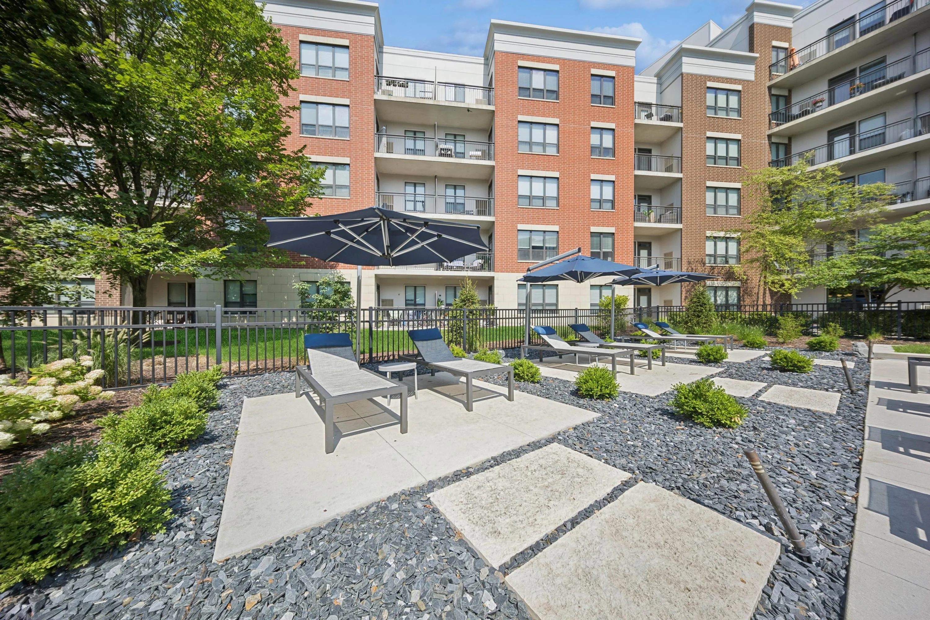 Chic urban apartment exterior with stylish lounge chairs and elegant umbrellas on a stone patio, surrounded by lush greenery.