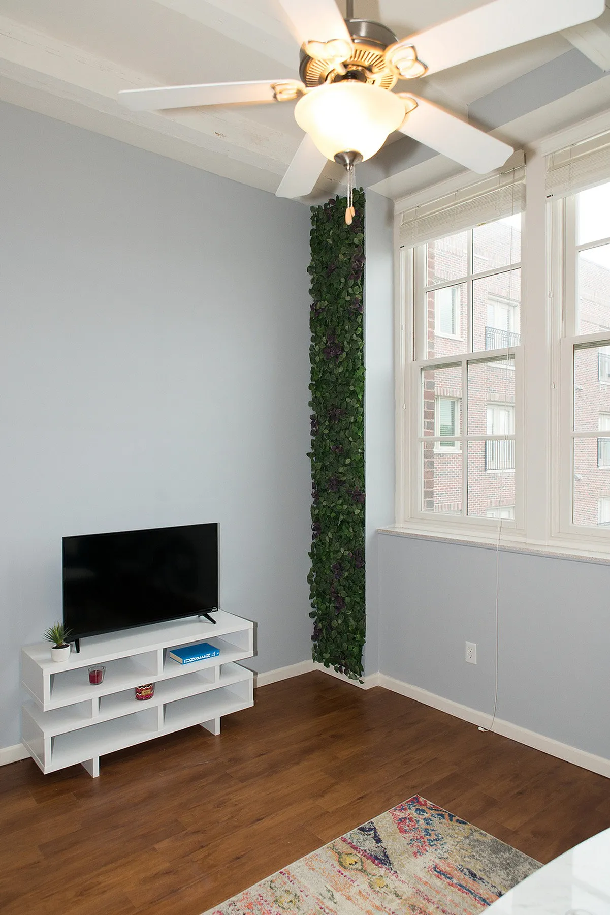 Bright living area with modern ceiling fan, stylish TV stand, and decorative greenery wall accent, featuring large windows for natural light.