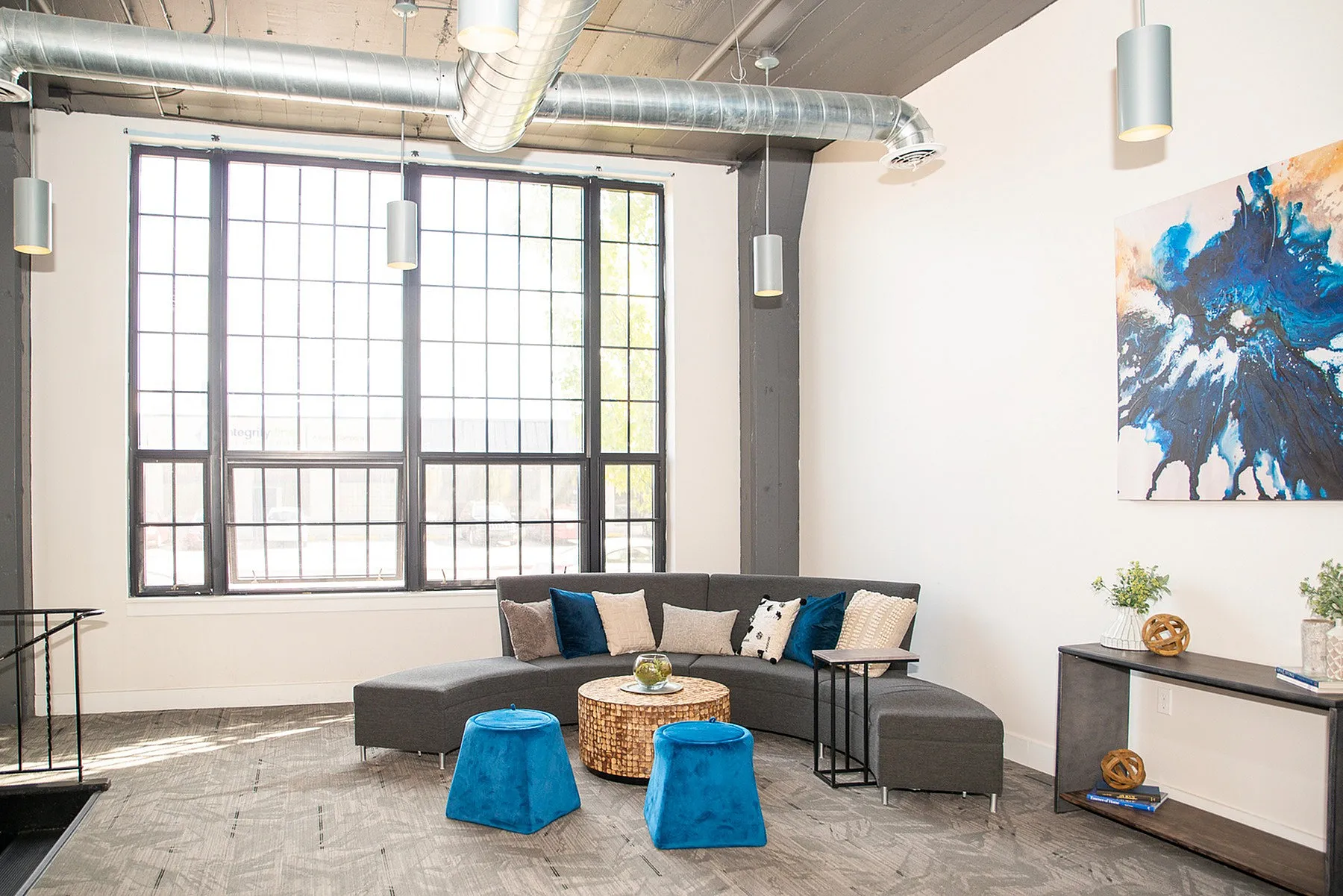 Bright, modern living room with industrial accents, featuring large windows, sleek gray sectional, and vibrant blue stools.