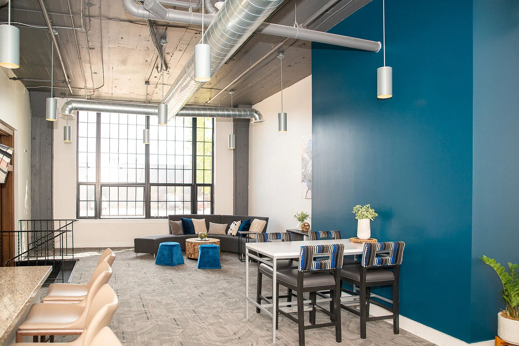 Modern loft-style living space with industrial aesthetic, featuring large windows, exposed ductwork, and a vibrant blue accent wall.