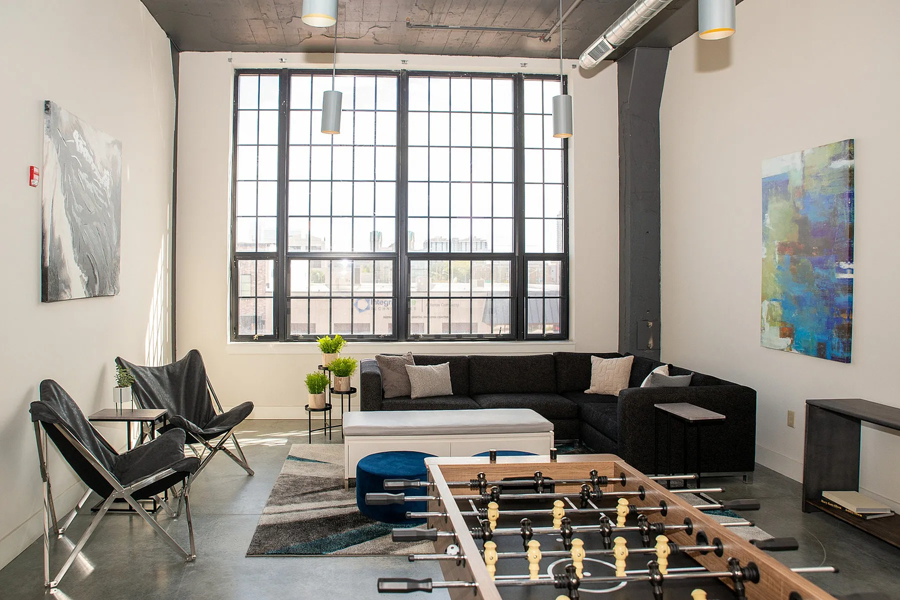 Chic urban living room with large industrial windows, modern furniture, and a stylish foosball table, perfect for entertaining.