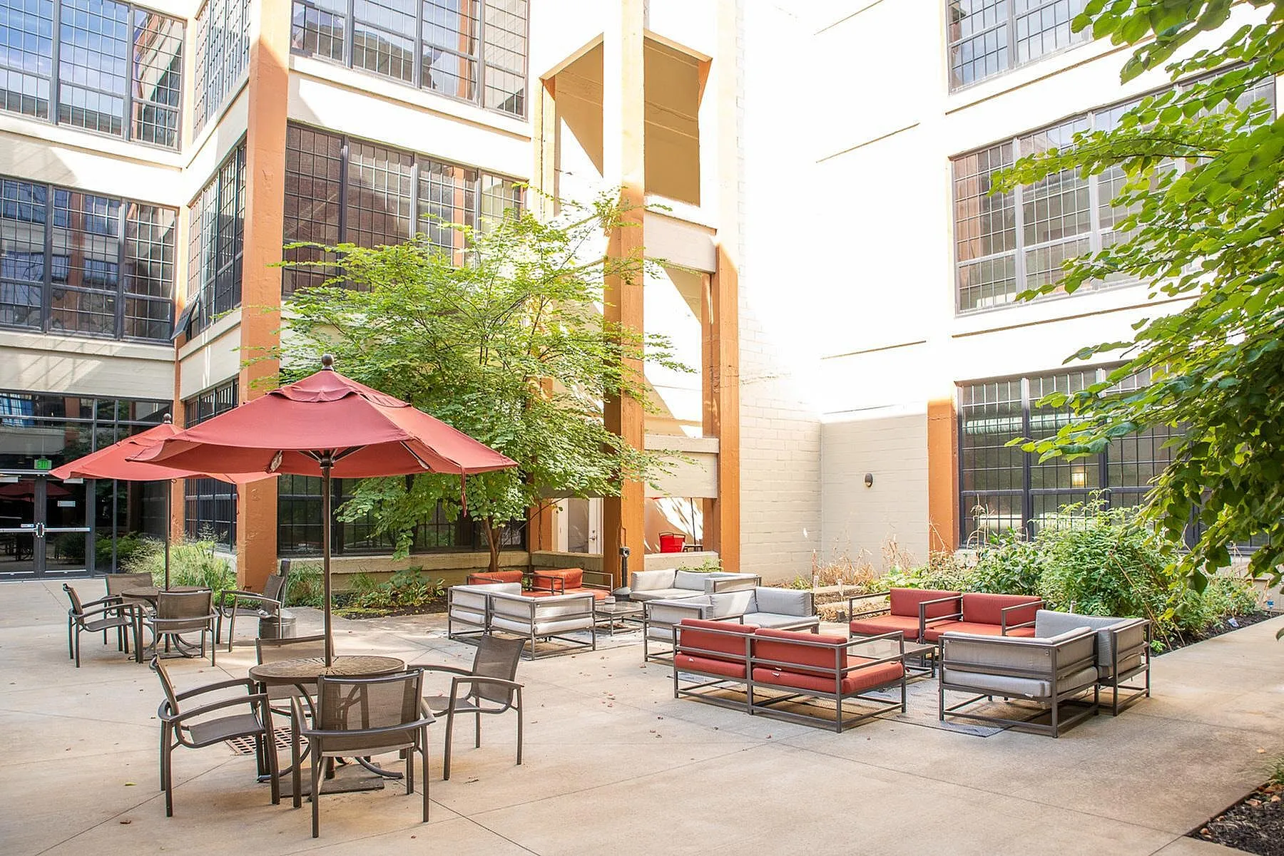 Welcoming courtyard of a modern apartment complex featuring stylish patio furniture, vibrant red umbrellas, lush greenery, and large industrial-style windows.