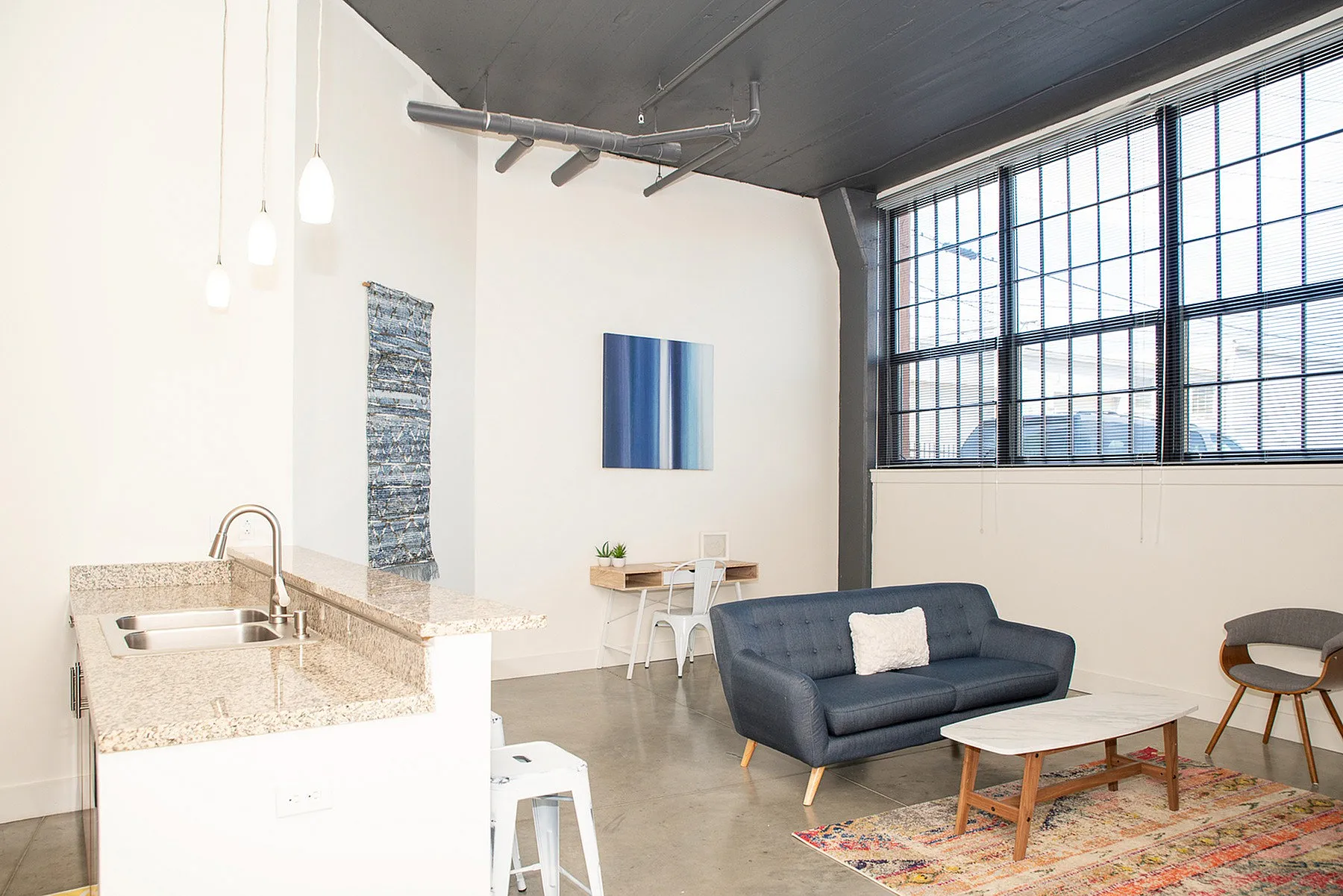 Modern open-plan living area featuring a cozy dark grey sofa, sleek kitchen with granite countertops, and large industrial-style windows for abundant natural light.