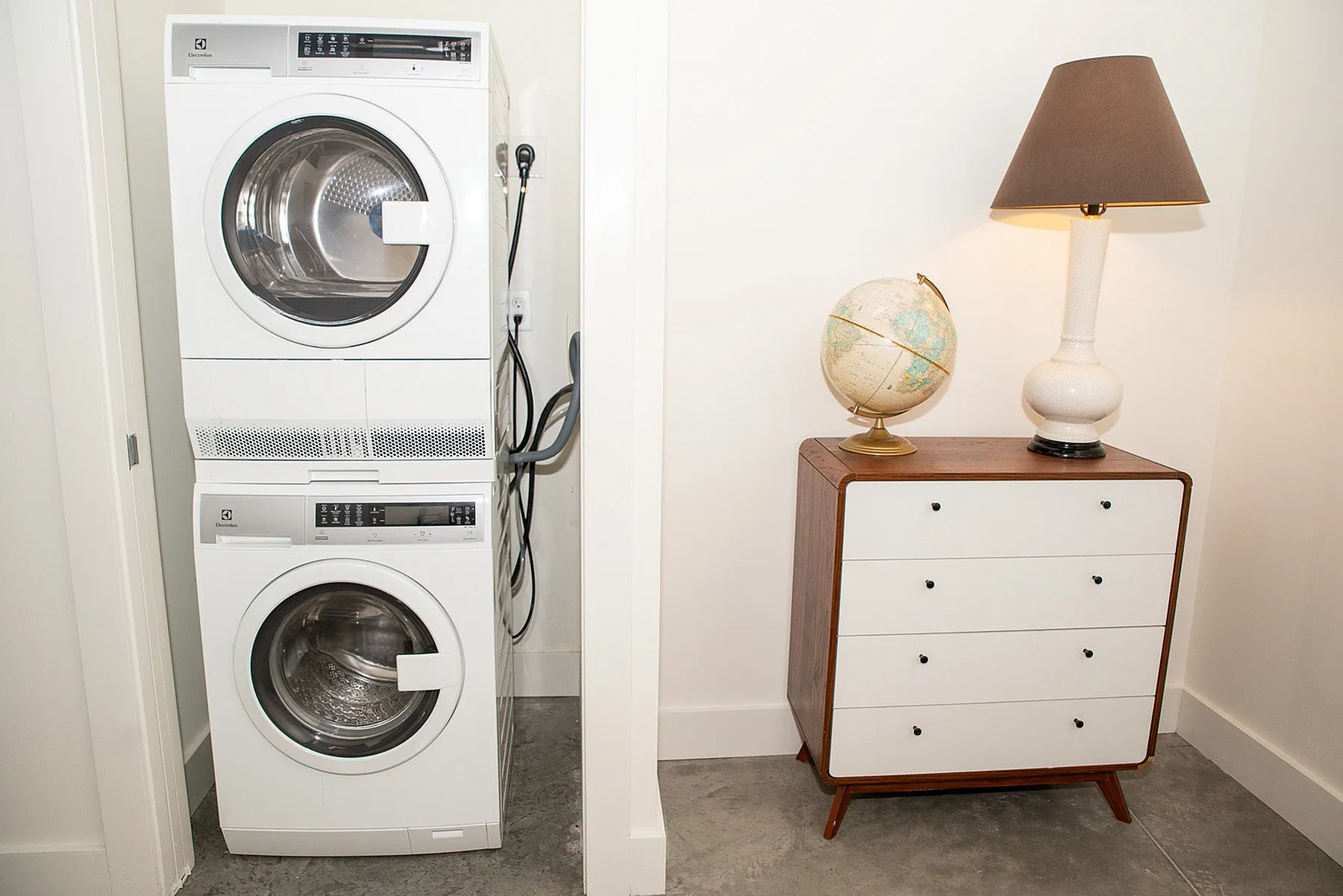 Contemporary laundry area with a sleek stacked washer and dryer, accompanied by a stylish mid-century dresser and vintage globe, creating a functional yet chic corner.