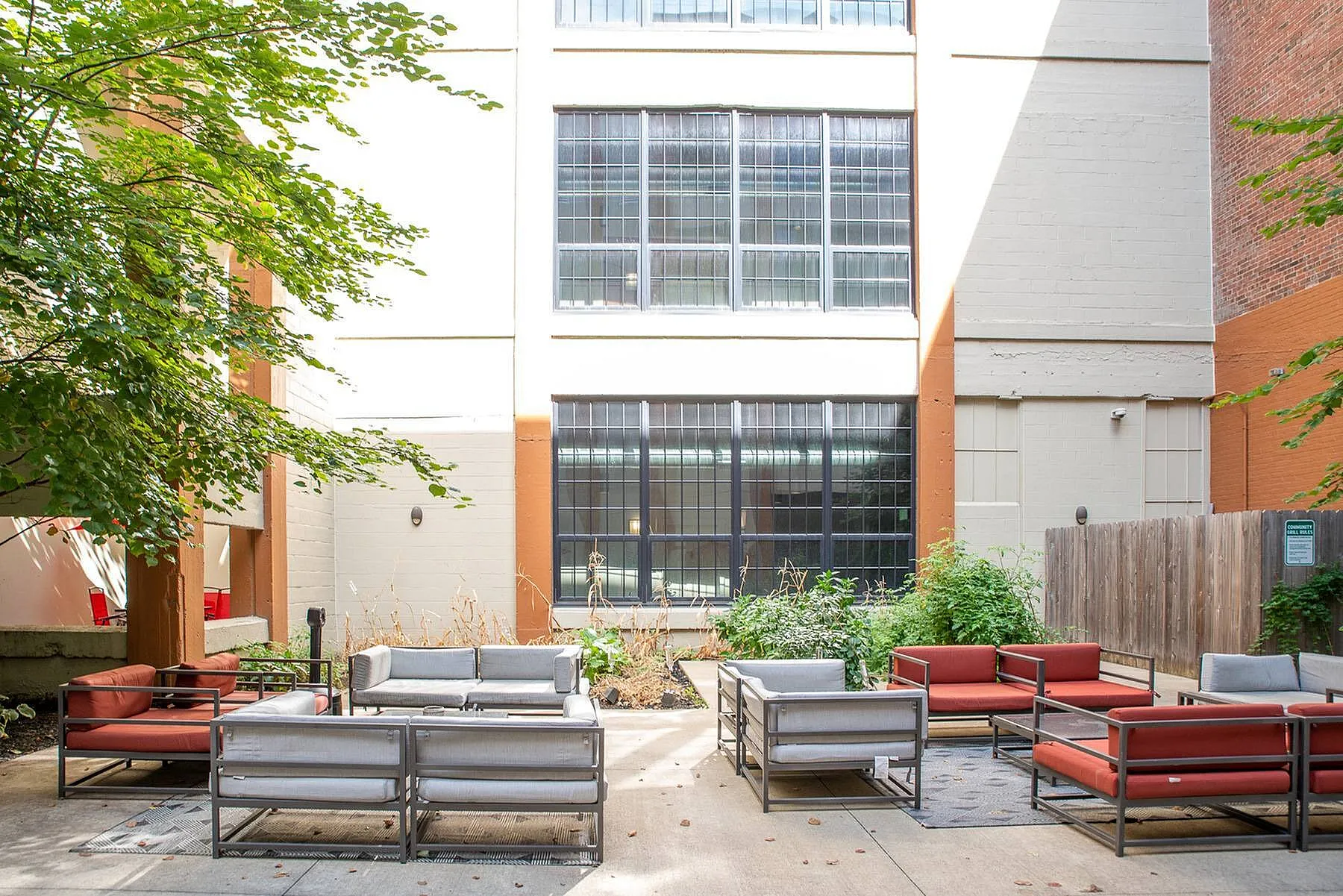 Charming urban courtyard with stylish patio seating, lush greenery, and large industrial-style windows, perfect for relaxation and social gatherings.