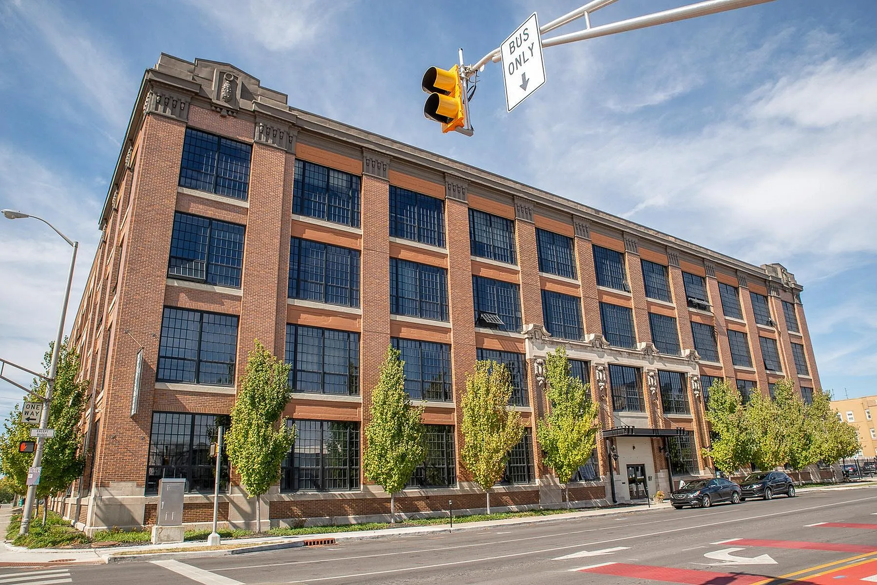 Elegant industrial-style apartment building with large windows and classic brick facade, surrounded by lush greenery on a sunny day.