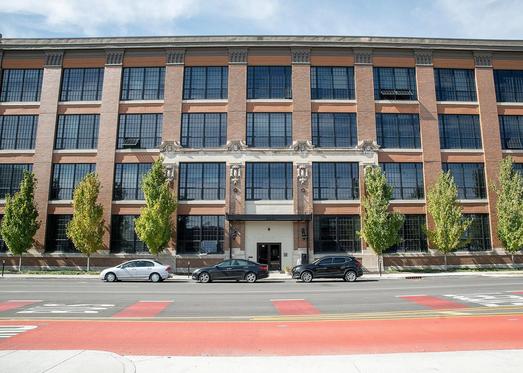 Charming industrial-style brick apartment building with large windows, surrounded by lush trees and set on a vibrant street.