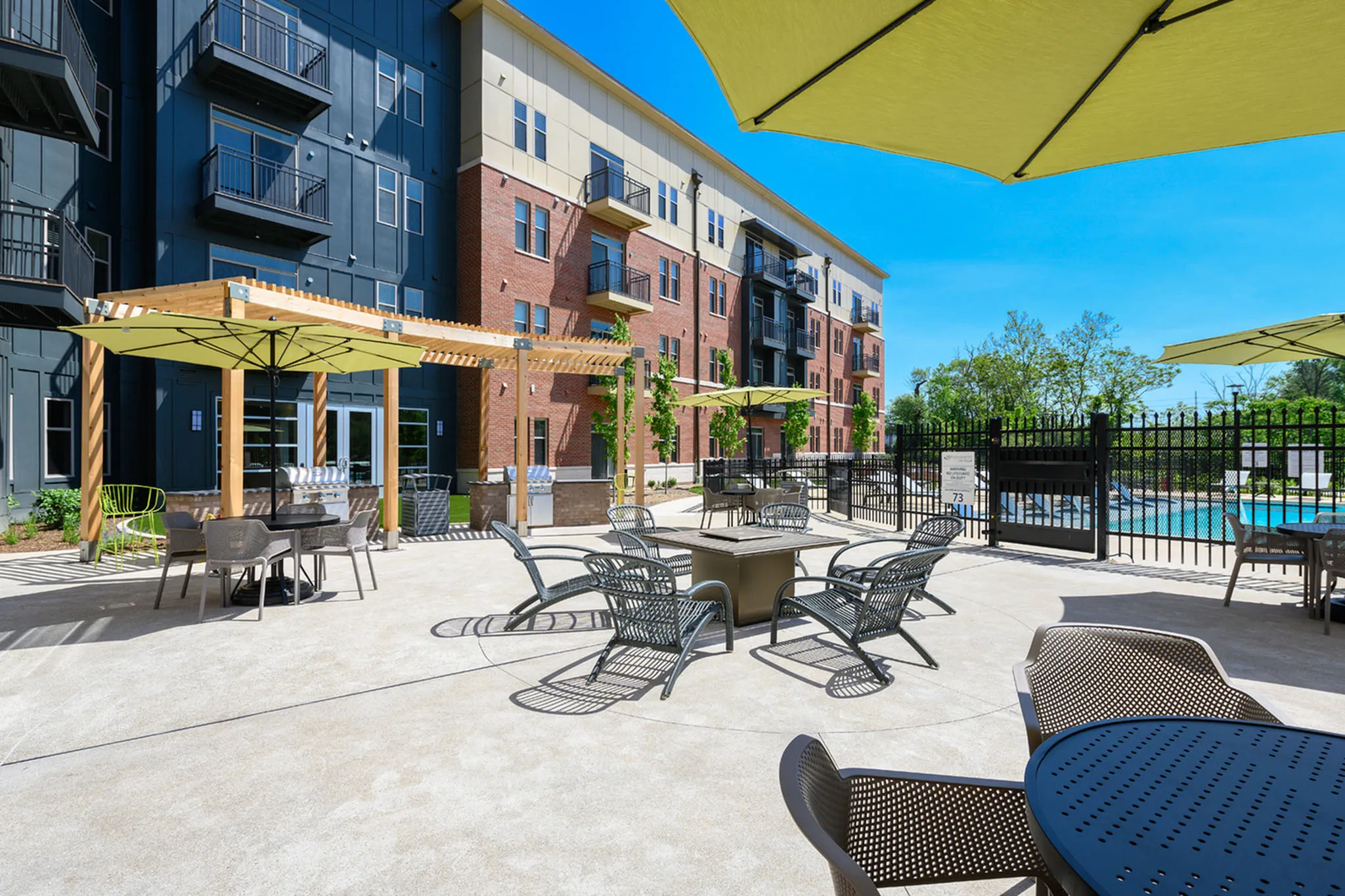 Modern apartment complex exterior with a spacious sunlit patio featuring chic outdoor seating, surrounded by lush greenery and vibrant blue skies.