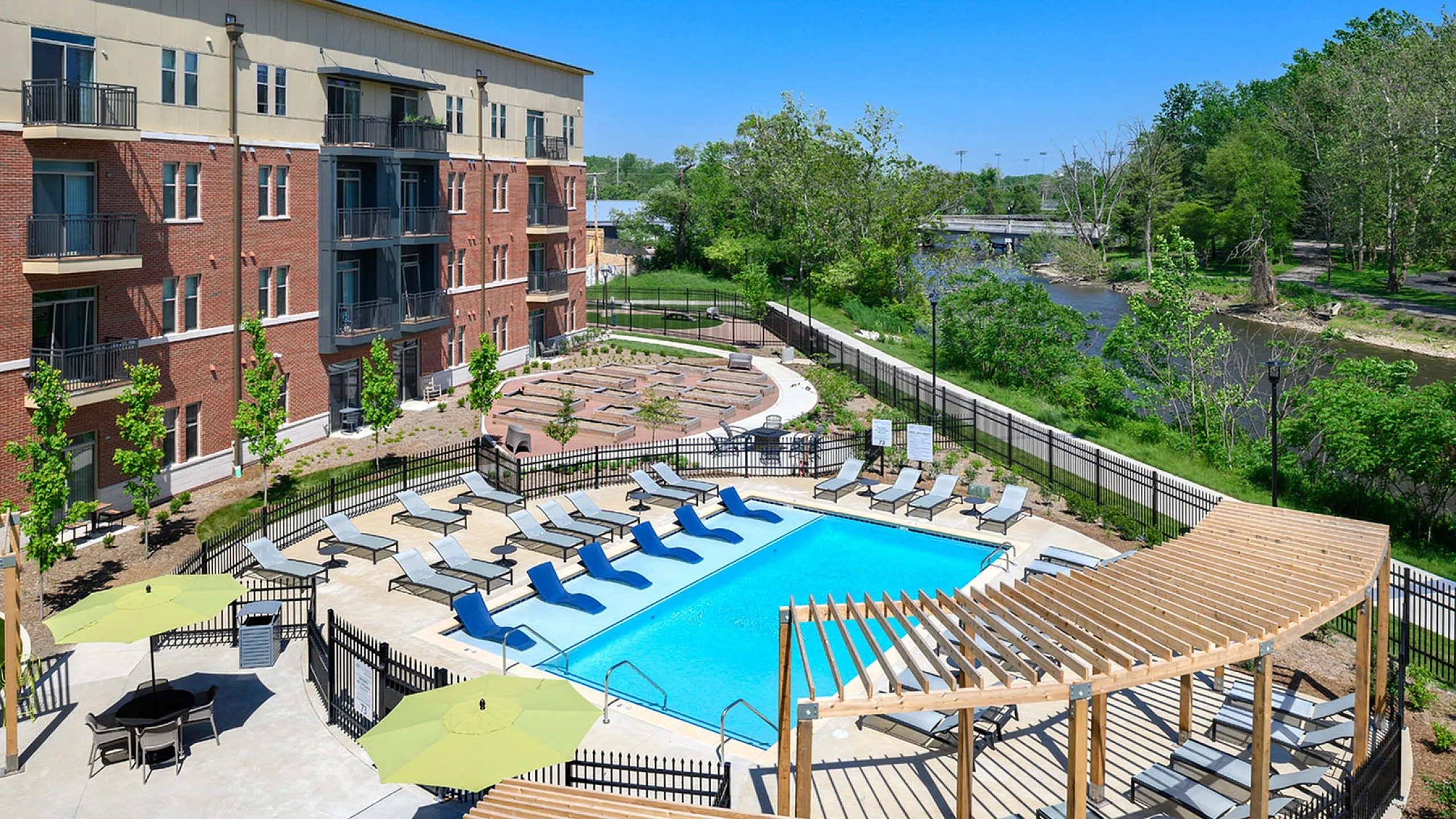 Elegant apartment complex featuring a modern swimming pool with ample sun loungers, surrounded by lush greenery and offering scenic river views.