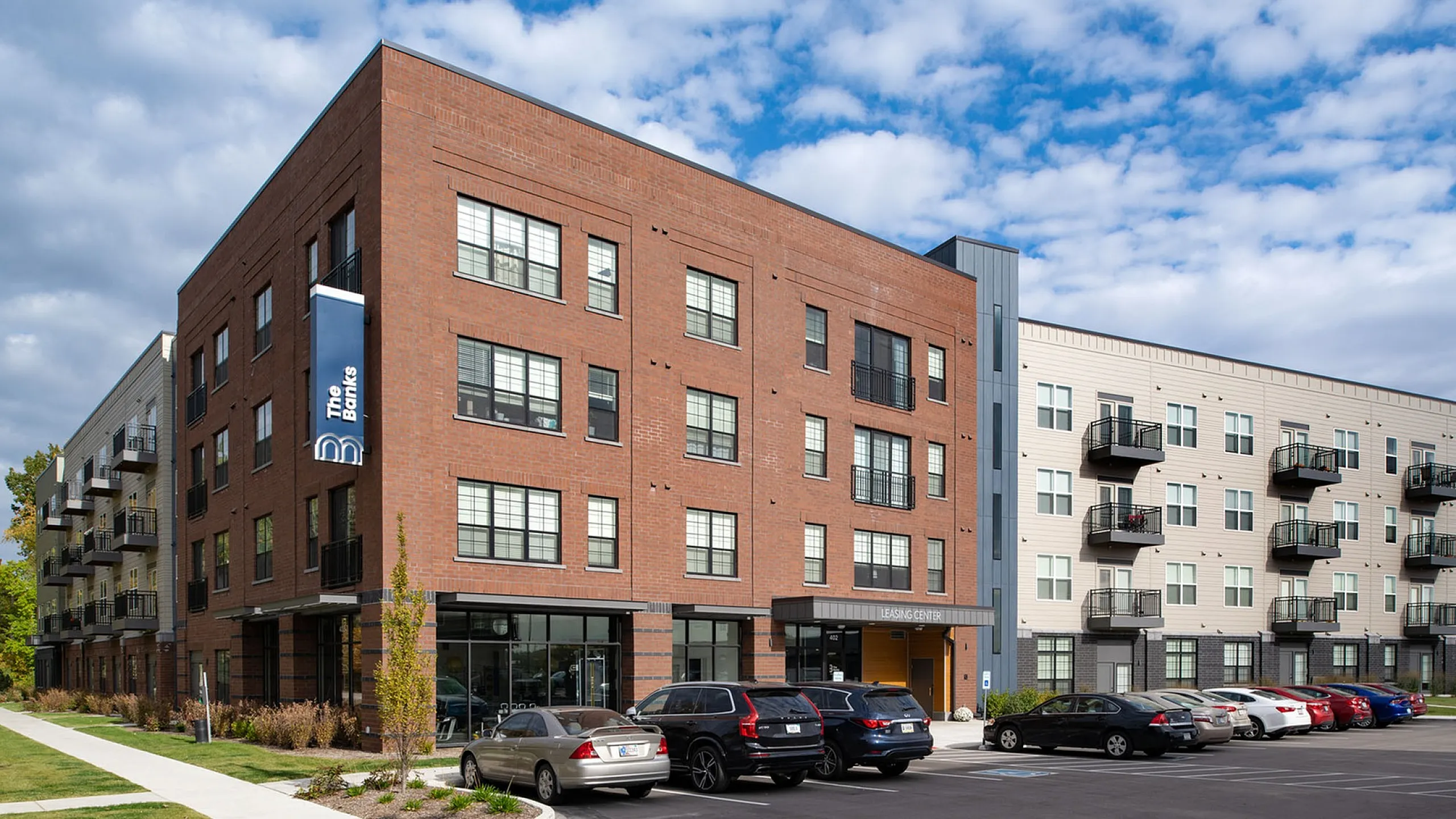 Modern apartment complex featuring a mix of brick and panel exterior, ample parking, and balconies for each unit, set against a vibrant blue sky.
