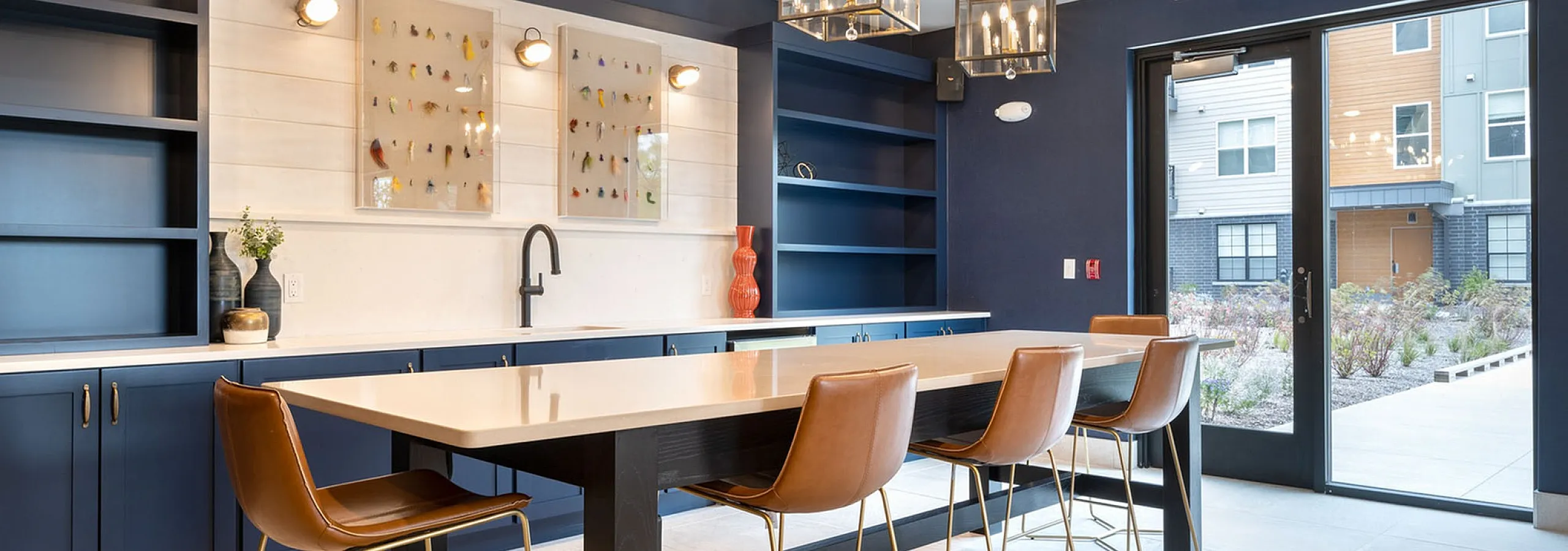 Modern communal dining area with elegant chandeliers, sleek navy cabinetry, and stylish leather chairs, showcasing contemporary design and natural light.
