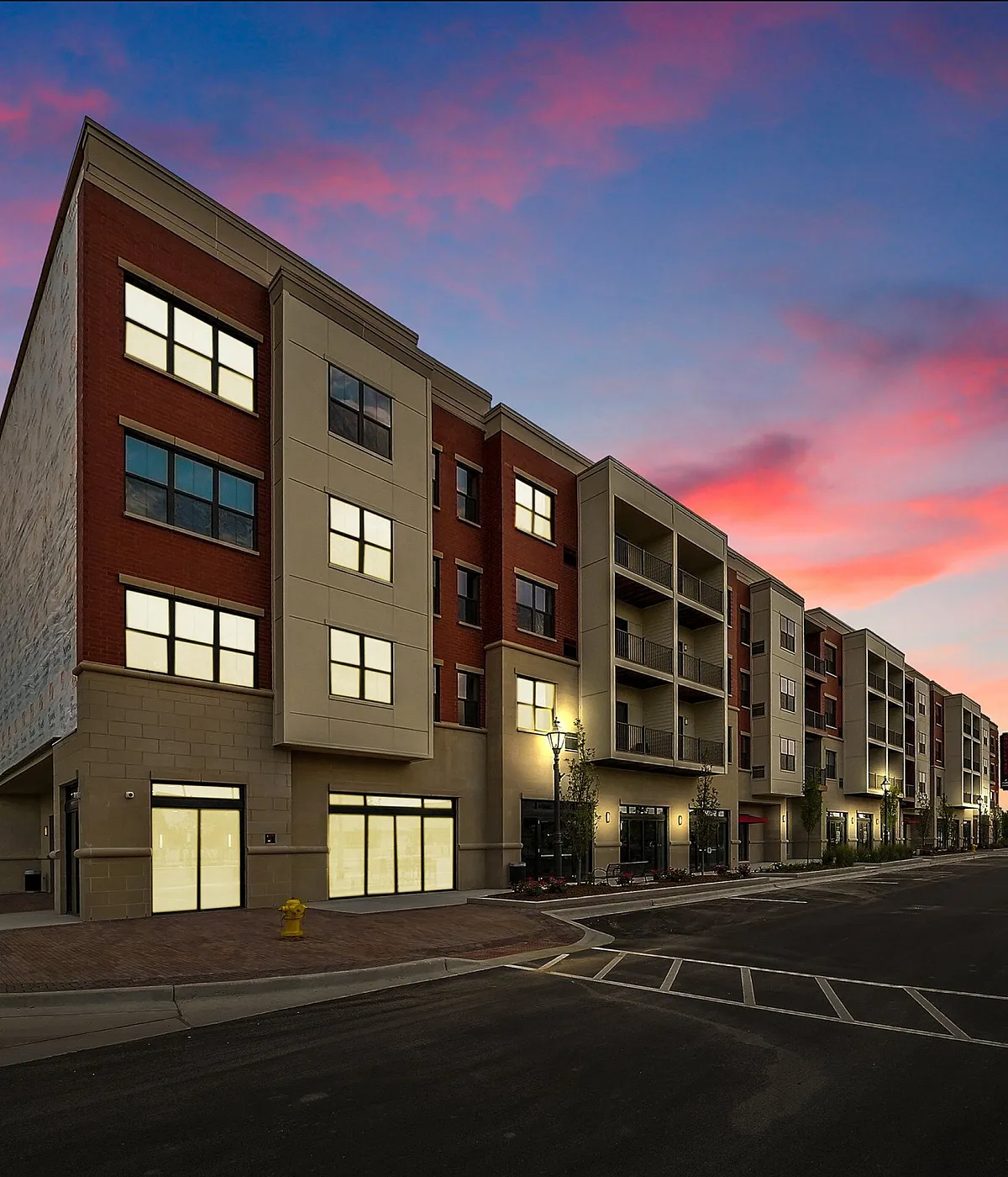 Modern apartment building at sunset with vibrant sky, featuring illuminated windows and a welcoming street entrance.