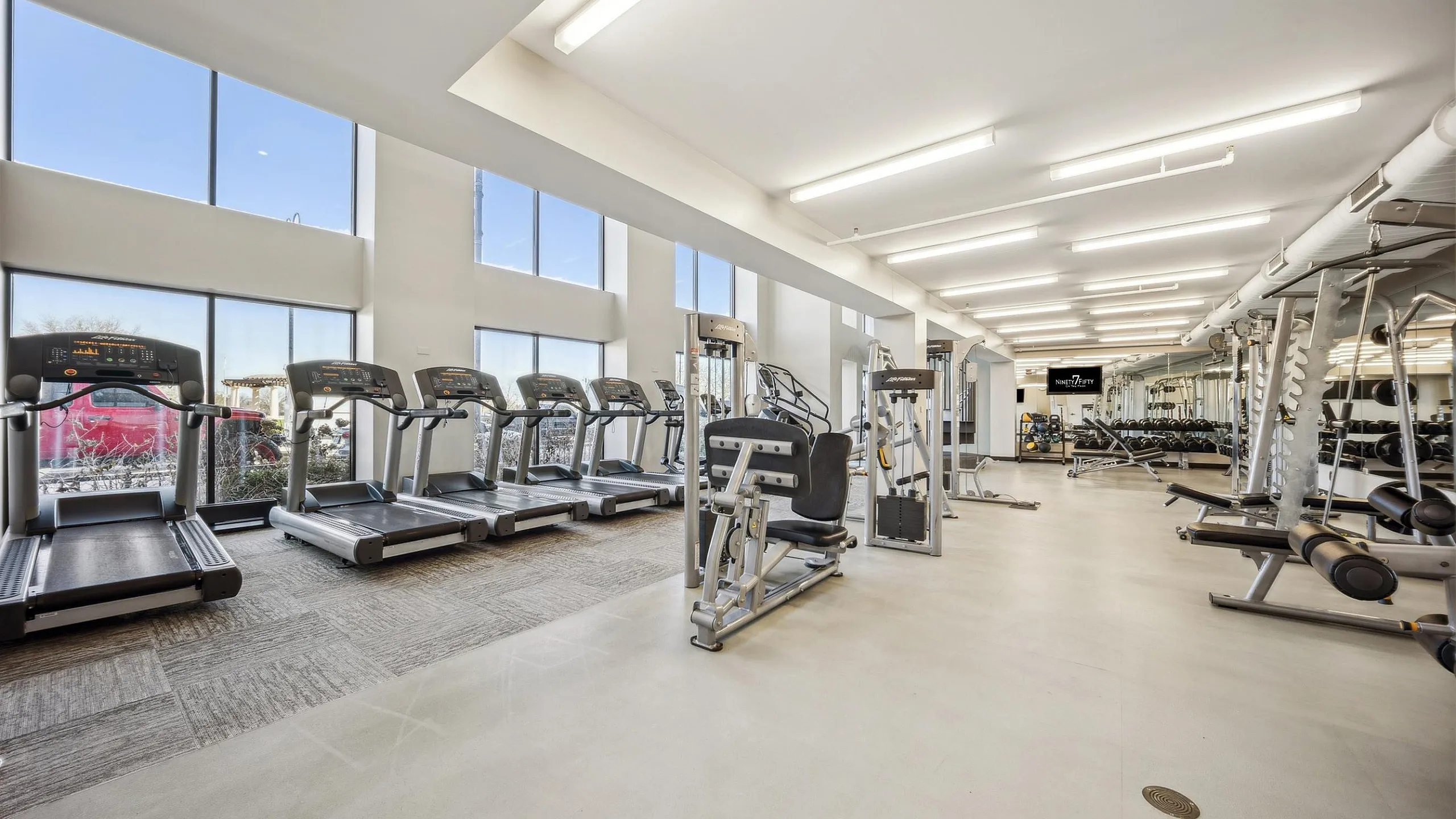 Bright and modern fitness center featuring a row of treadmills, workout equipment, and expansive windows.
