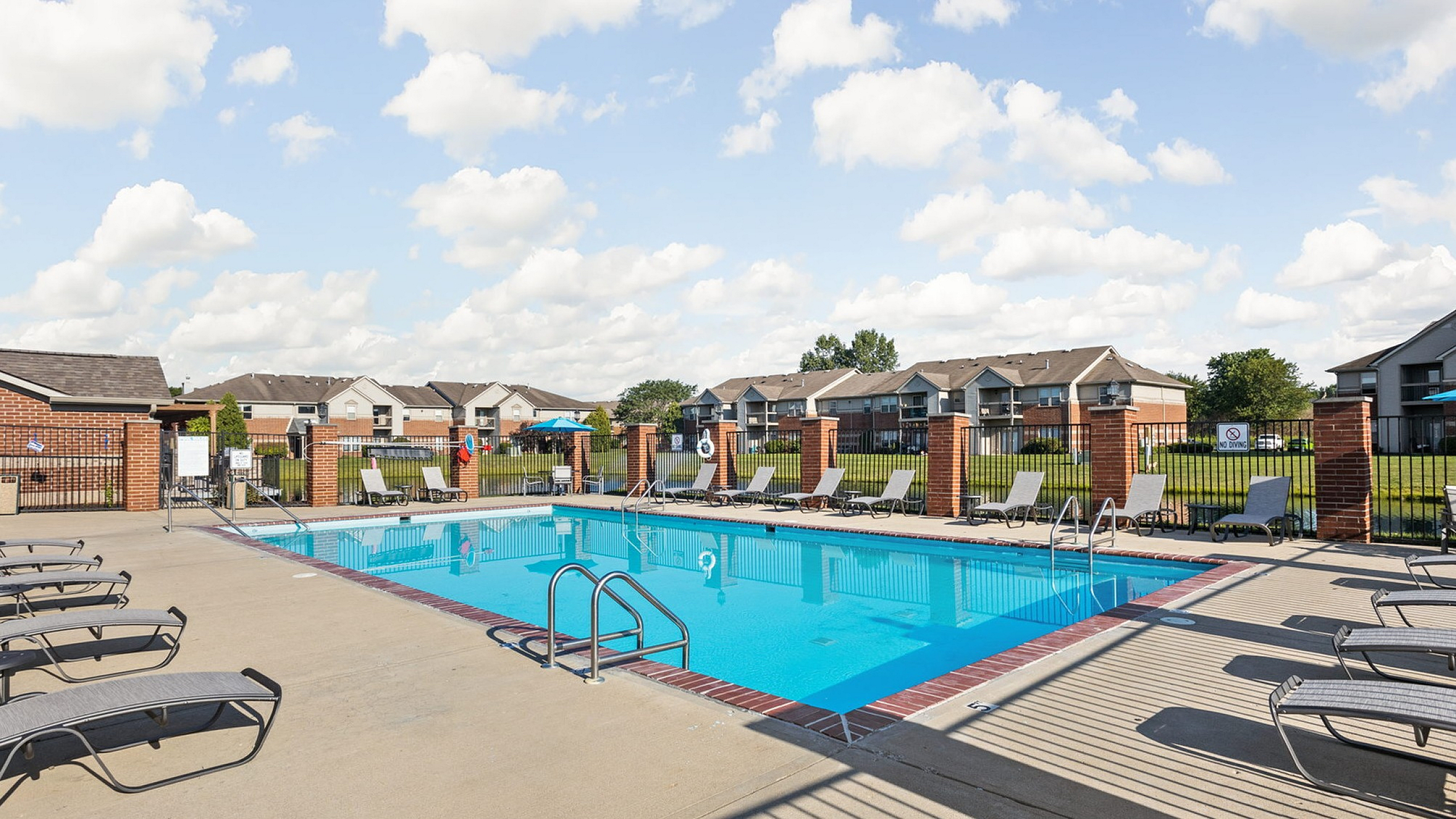 Inviting apartment community with a sparkling swimming pool surrounded by comfortable lounge chairs and stylish brick fencing under a bright, clear sky.