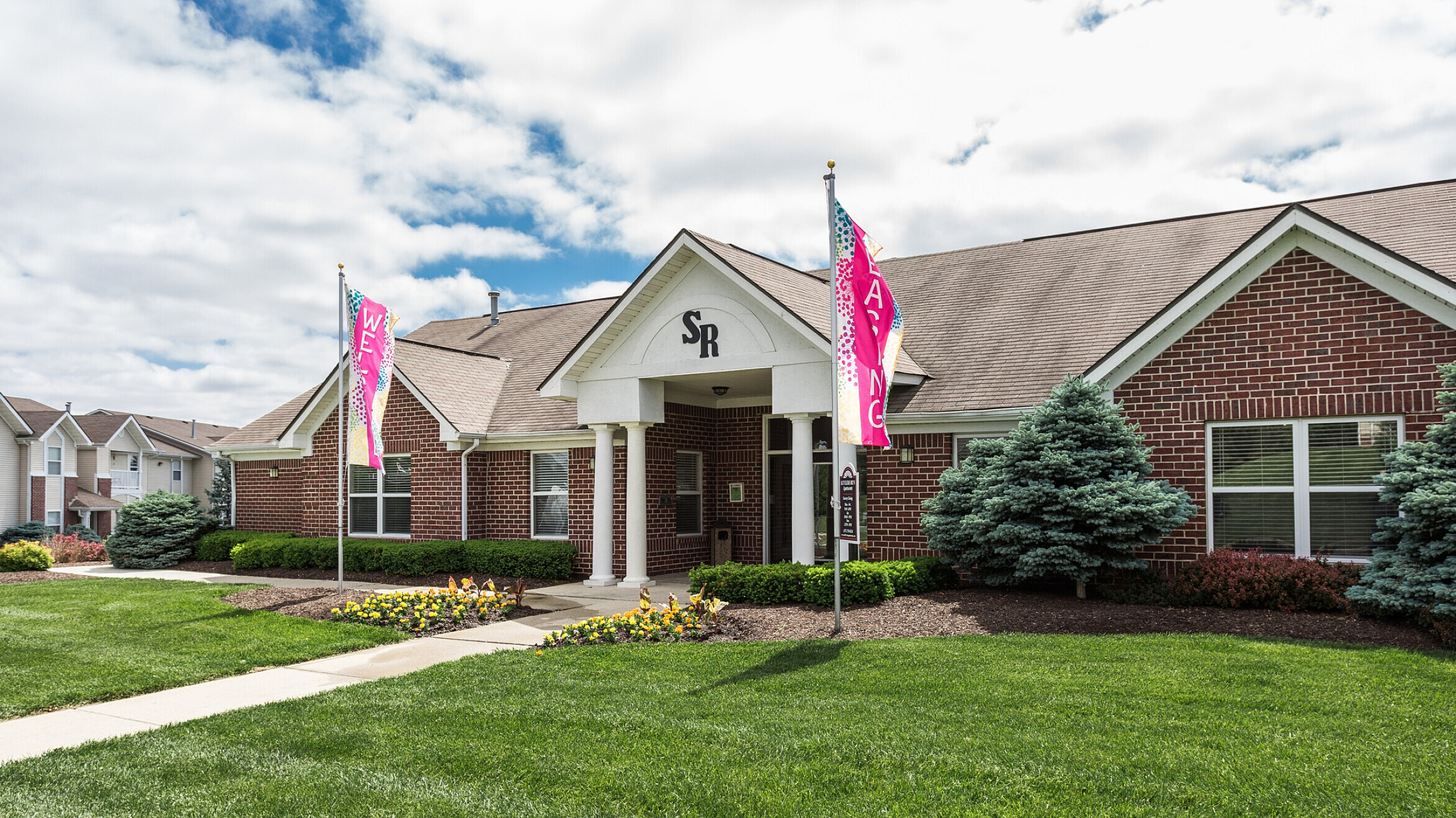 Charming brick clubhouse with well-maintained gardens and vibrant flags, offering a warm welcome to a thriving apartment community.