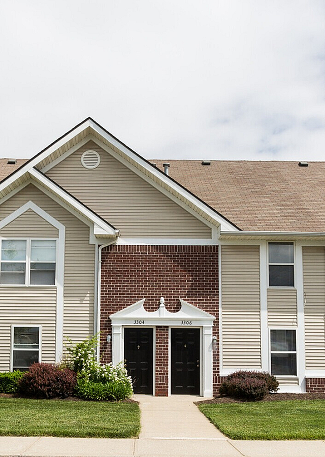 Charming townhouse complex with classic brick and siding exterior, surrounded by lush greenery and spacious parking, under a bright blue sky.