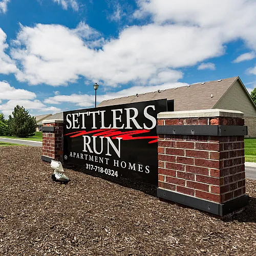 Inviting entrance to Settlers Run Apartment Homes on a sunny day, featuring a neatly landscaped pathway and vibrant community sign.