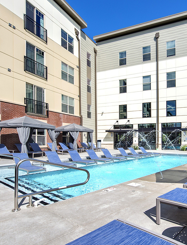 Luxury apartment complex featuring a stunning outdoor pool area with elegant loungers, shaded cabanas, and vibrant water features under a clear blue sky.