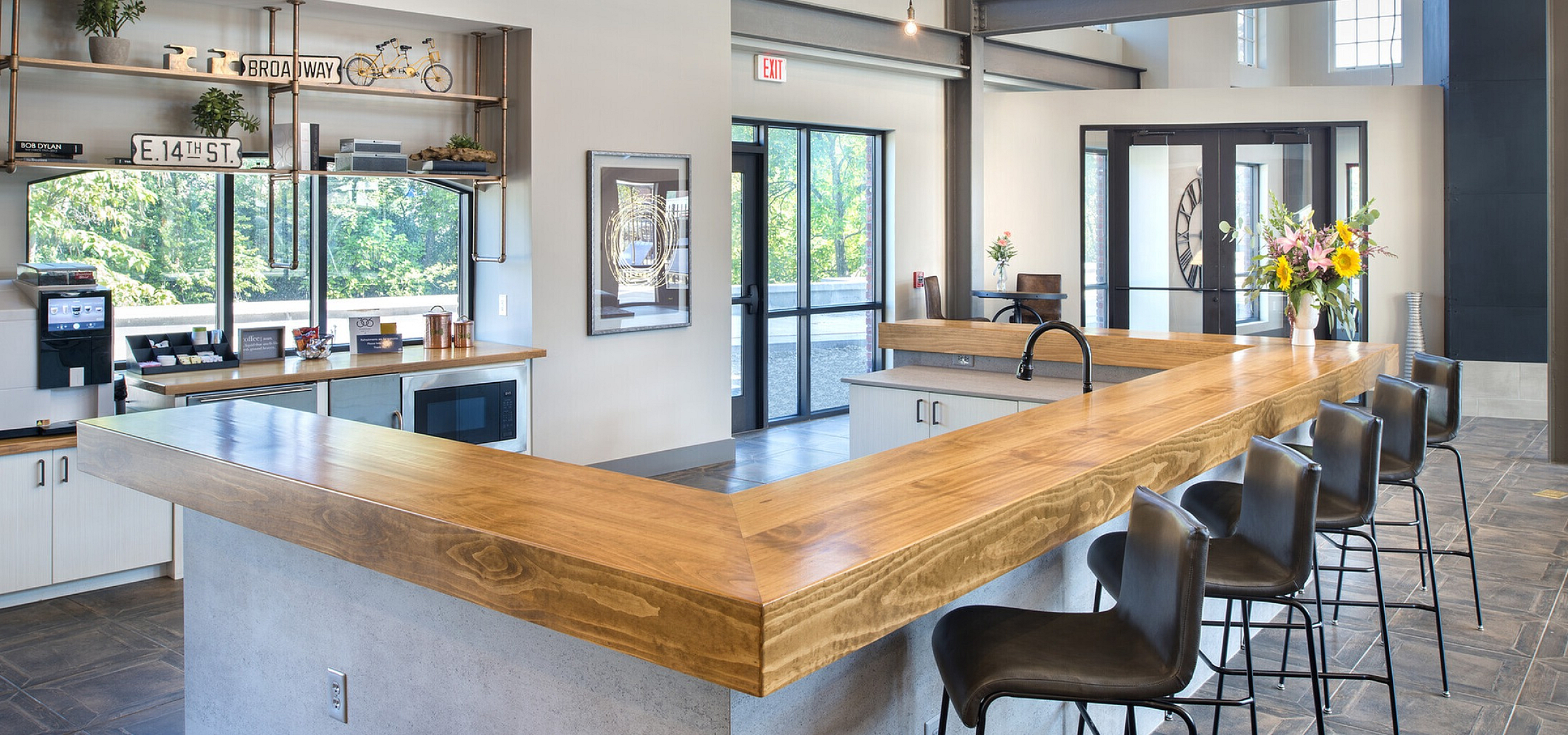 Modern industrial-style kitchen with high ceilings, large windows, and a sleek wooden breakfast bar surrounded by stylish bar stools, offering a bright and spacious atmosphere.