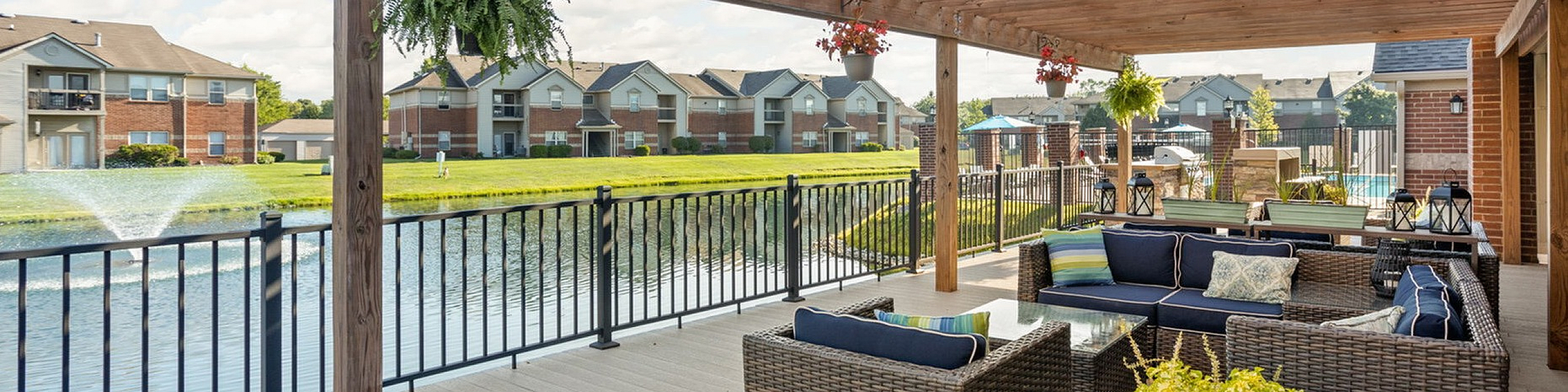 Charming apartment deck with cozy seating under a wooden pergola, overlooking a serene pond with a water fountain and lush greenery.