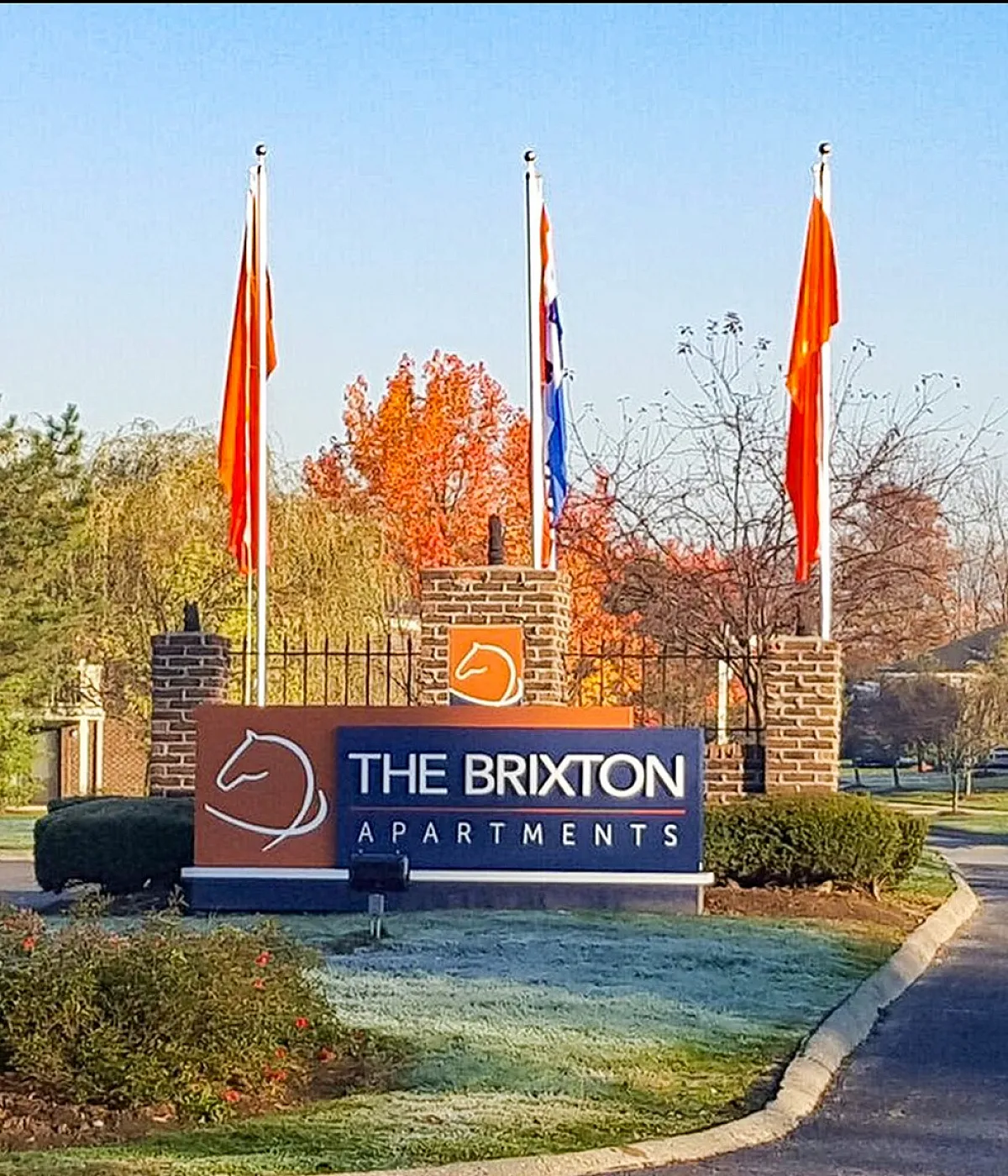 Charming entrance to The Brixton Apartments with vibrant flag display, surrounded by lush greenery and autumn trees.