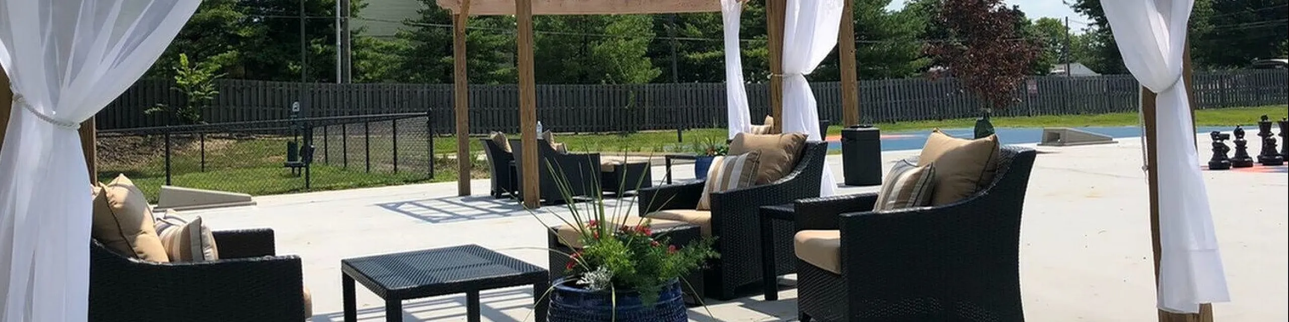 Elegant outdoor seating area under a stylish pergola with flowing white curtains, lush greenery, and comfortable wicker furniture.