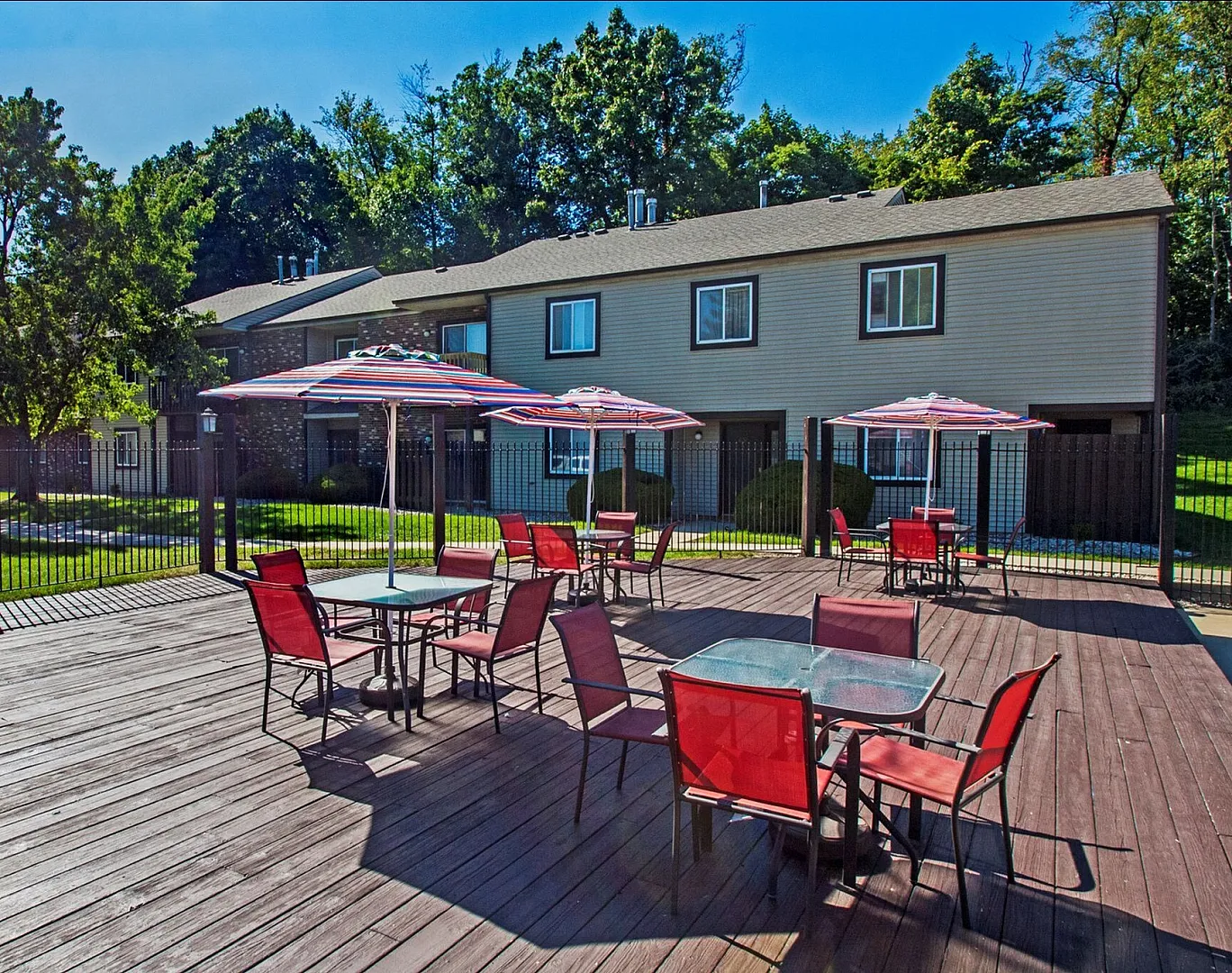 Charming outdoor patio with vibrant red chairs and striped umbrellas, perfect for relaxing under the sun in a lush, green apartment complex.