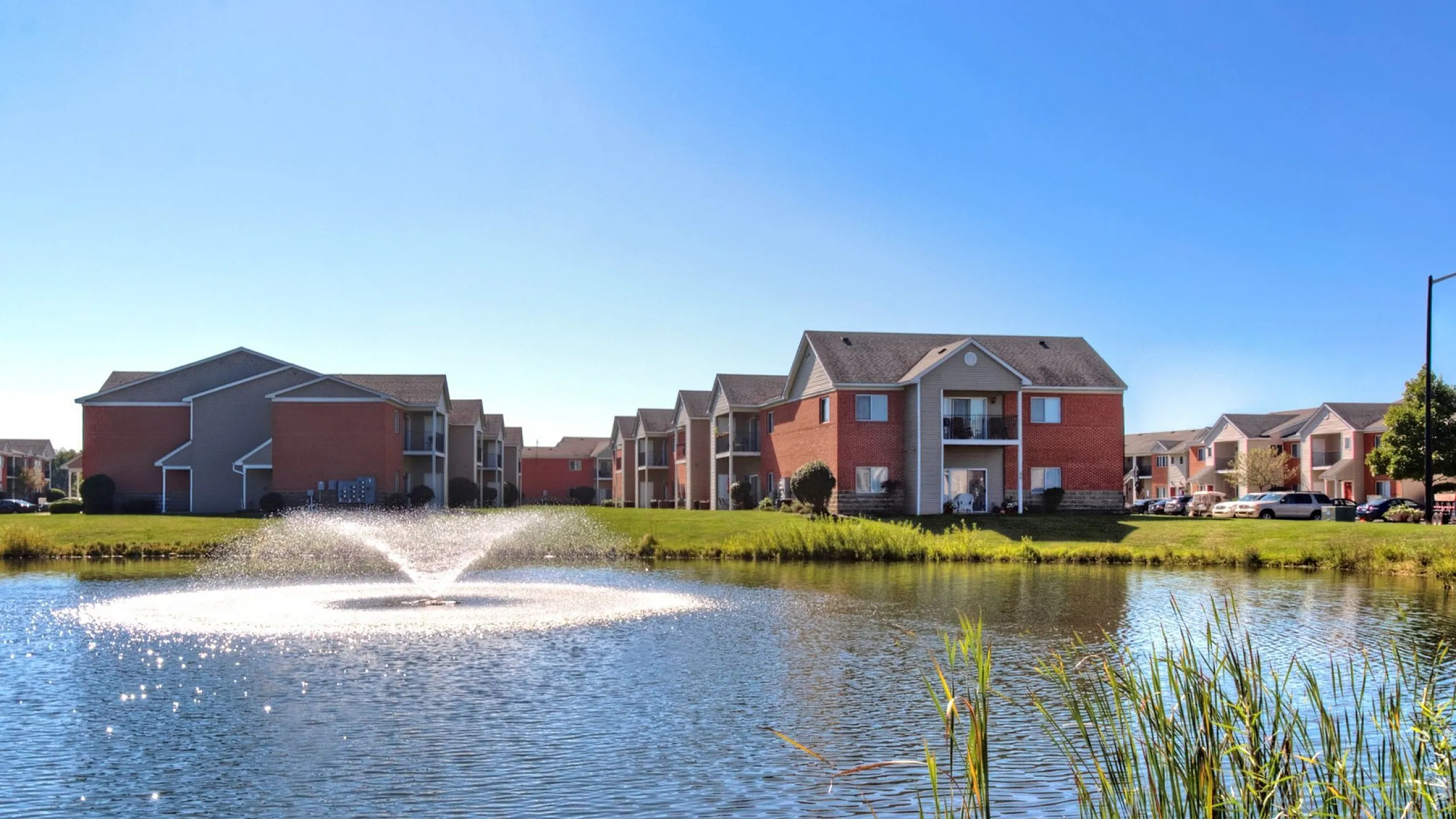Charming apartment complex with picturesque pond view, featuring a scenic water fountain and lush greenery.