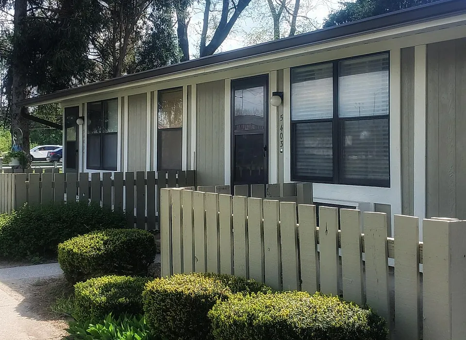 Charming single-story apartment with a modern exterior, surrounded by lush greenery and a quaint wooden fence, offering serene living.
