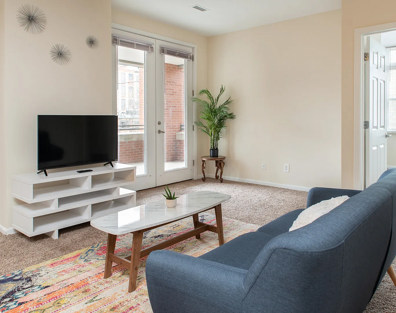 Bright and inviting living room with stylish decor, modern TV unit, and cozy seating area opening to a sunlit balcony with indoor plant accent.