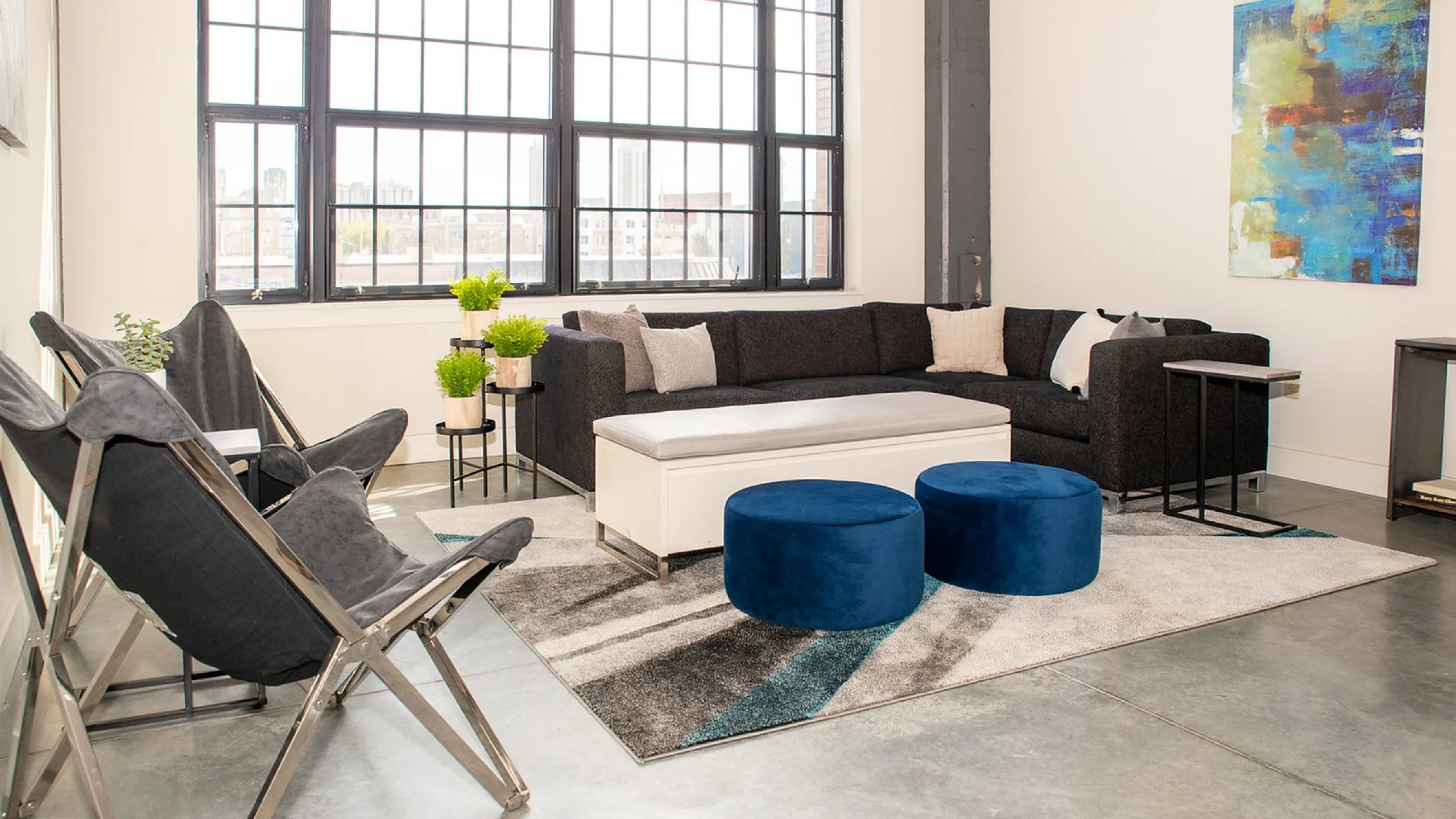 Bright and modern living space featuring a sleek black sectional, plush white ottoman, and vibrant blue stools beneath large industrial-style windows.