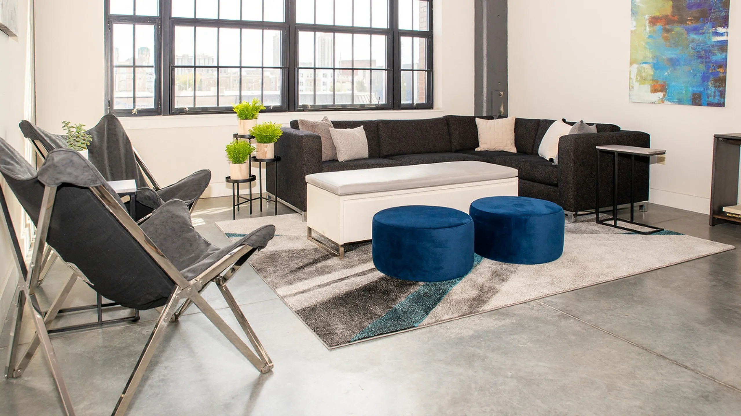 Chic modern living room with industrial-style windows, plush sectional, vibrant artwork, and stylish blue ottomans on a sleek rug.