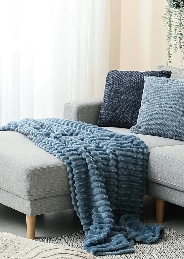 Chic living room corner with stylish grey sofa, plush blue throw, and lush green potted plant beside sheer curtains.