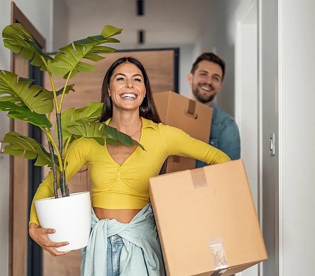 Happy couple moving into a bright modern apartment, carrying boxes and a green potted plant.