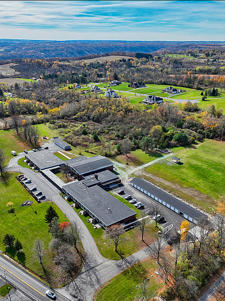 Scenic aerial view of a spacious countryside property with modern buildings surrounded by lush greenery and rolling hills under a clear blue sky.
