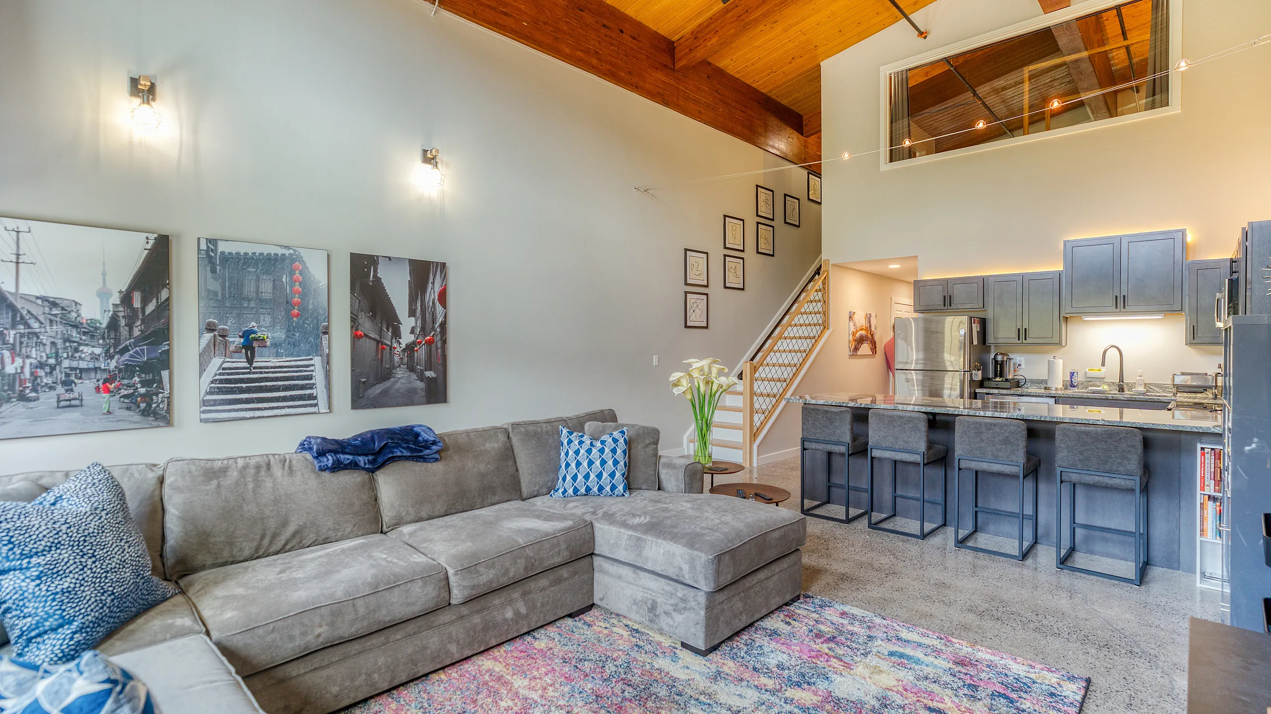 Contemporary open-plan living room with plush sectional sofa, vibrant rug, and modern kitchen with sleek bar seating under a warm wooden ceiling.