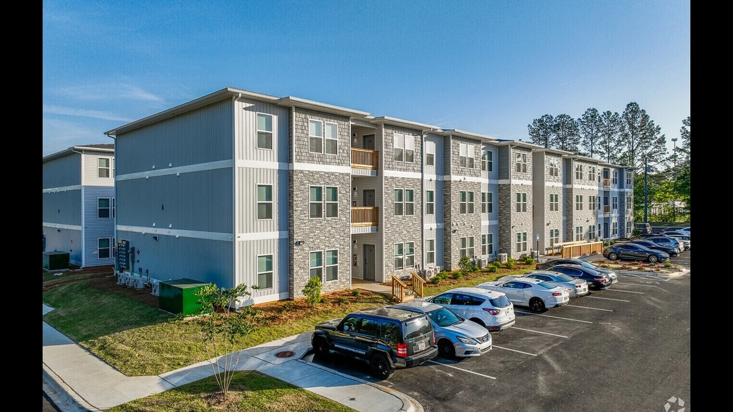 Modern apartment complex with sleek exterior design, ample parking, and lush green surroundings under a clear blue sky.