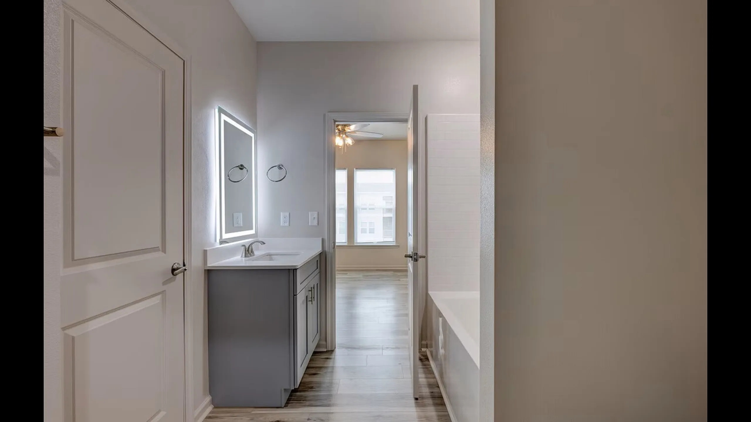 Bright and modern bathroom featuring a sleek vanity with an illuminated mirror, leading to a sunlit bedroom area.