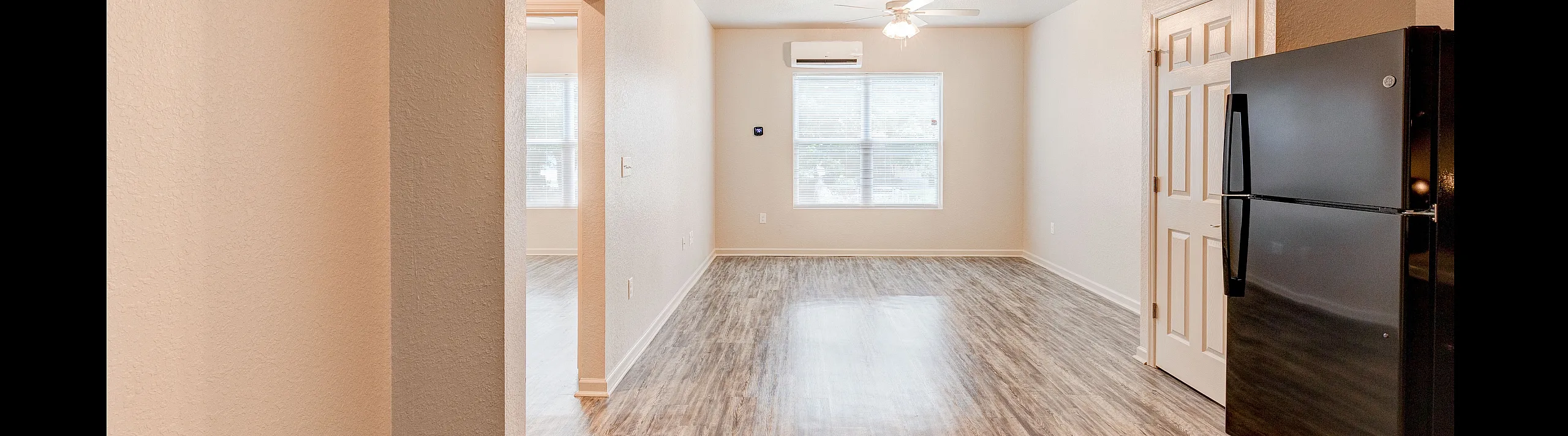 Bright and airy apartment interior featuring an open living area with modern flooring, large window, ceiling fan, and sleek black refrigerator.