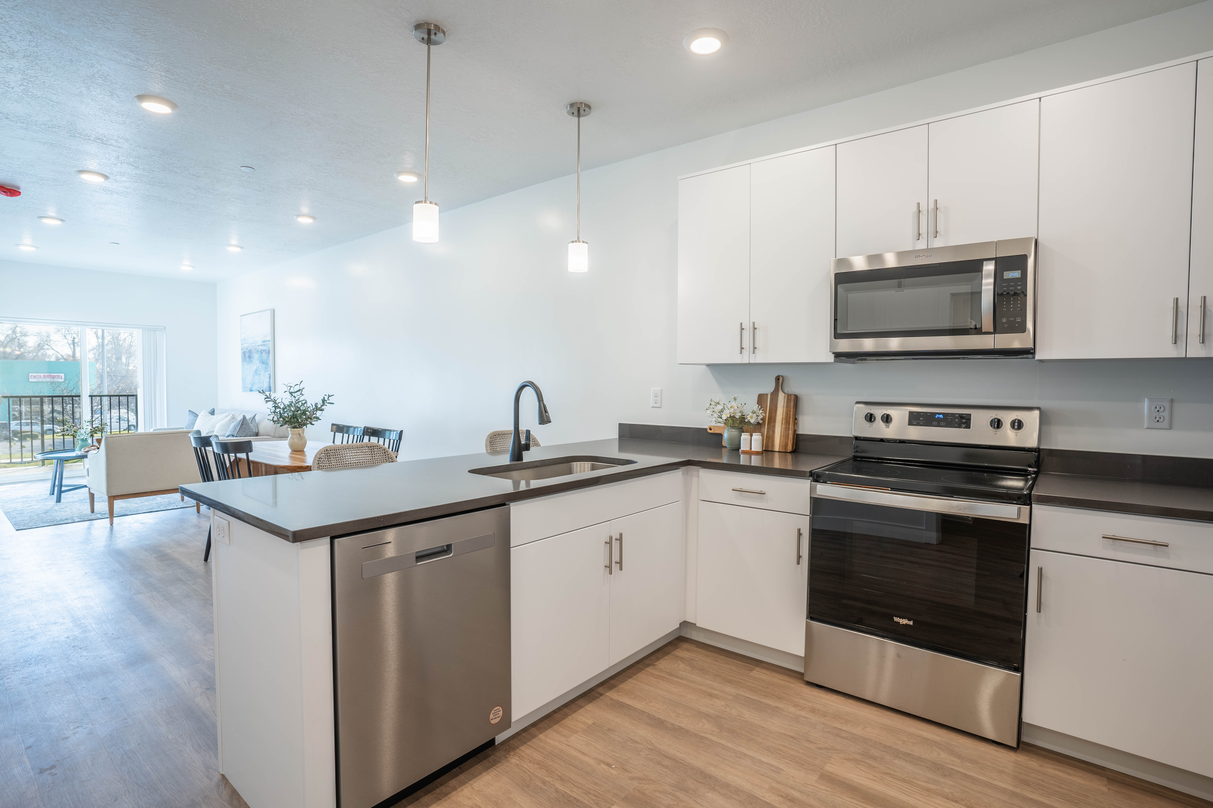 Modern open-plan kitchen with sleek white cabinetry, stainless steel appliances, and warm wooden floors leading to a bright living and dining area with large windows.