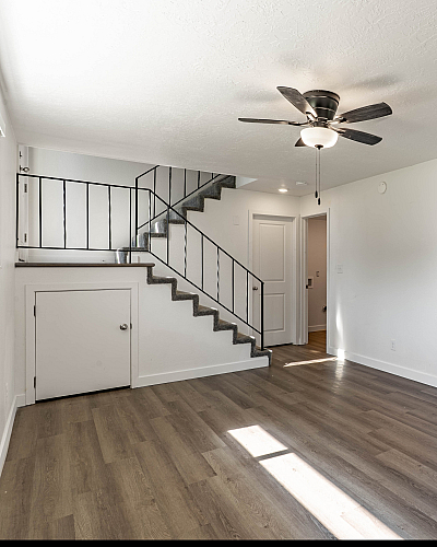 Bright open-concept living space with modern ceiling fan, sleek hardwood floors, and stairway leading to upper level, adjacent to a cozy kitchen with stylish cabinetry.