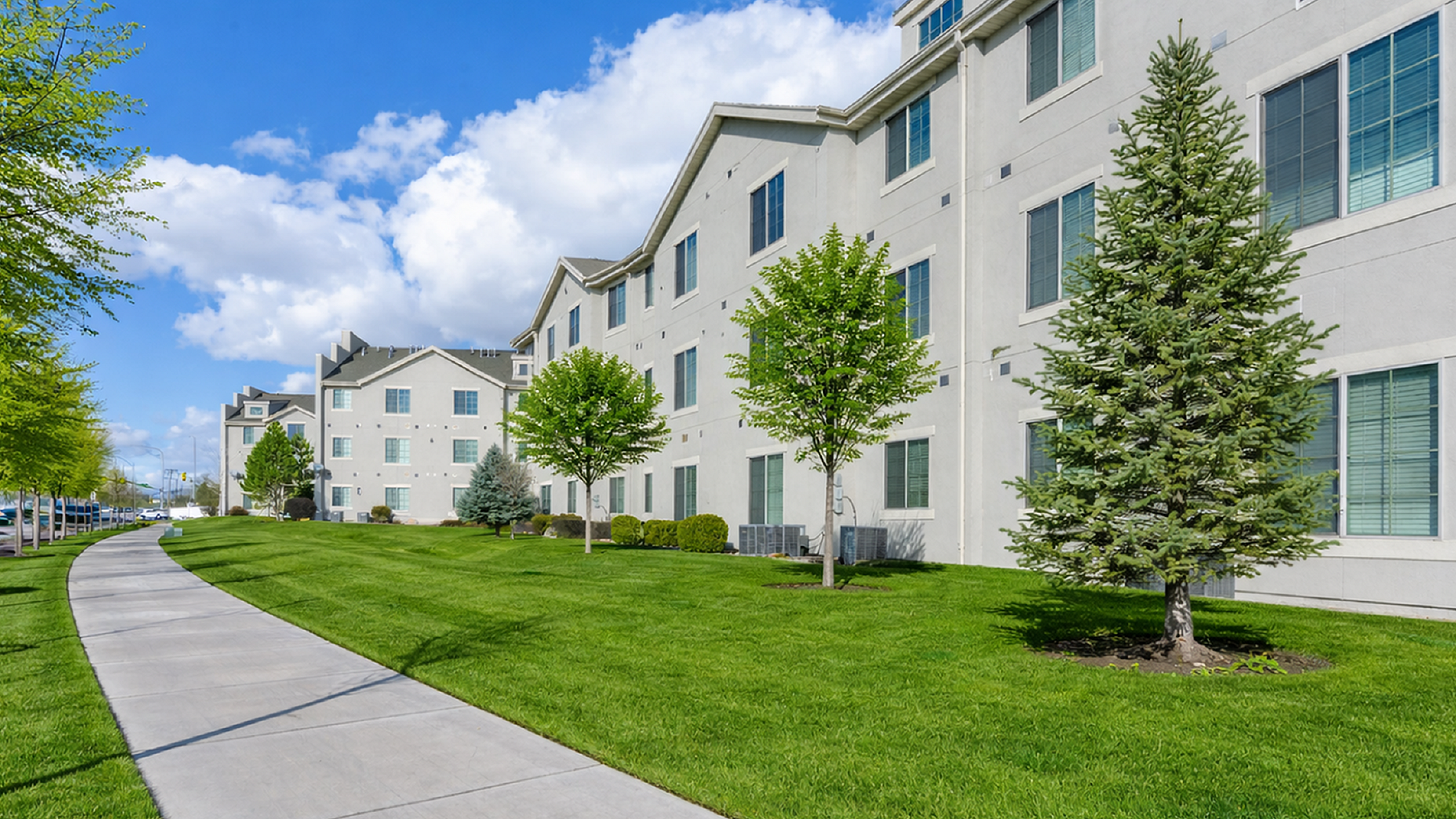 Modern apartment complex with manicured lawns and scenic walkways, set against a backdrop of vibrant greenery and clear blue skies.